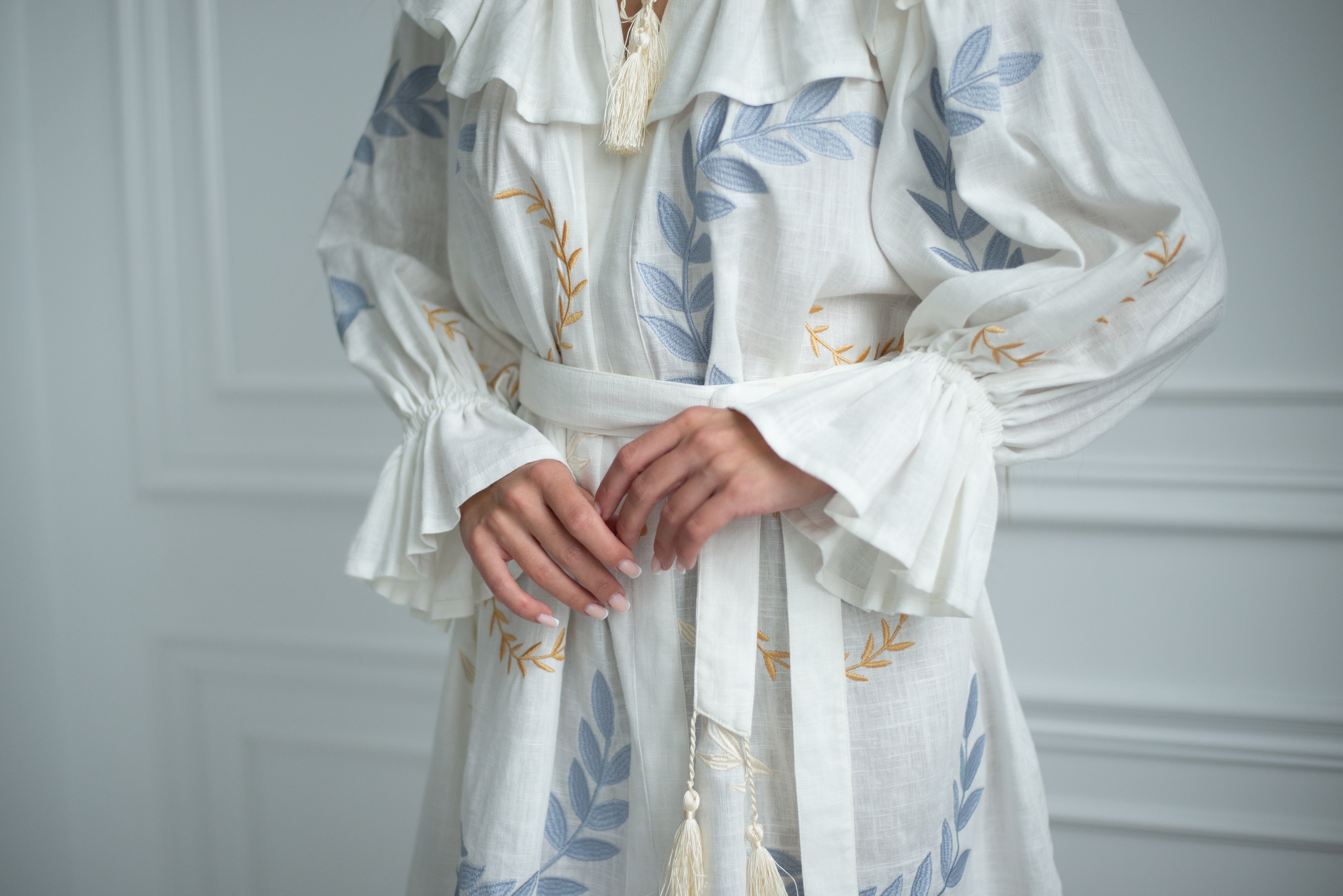 A close-up of a person wearing a white linen-style long-sleeve robe or dress with blue leaf embroidery, gold leaf embroidery, and a matching white tie belt.