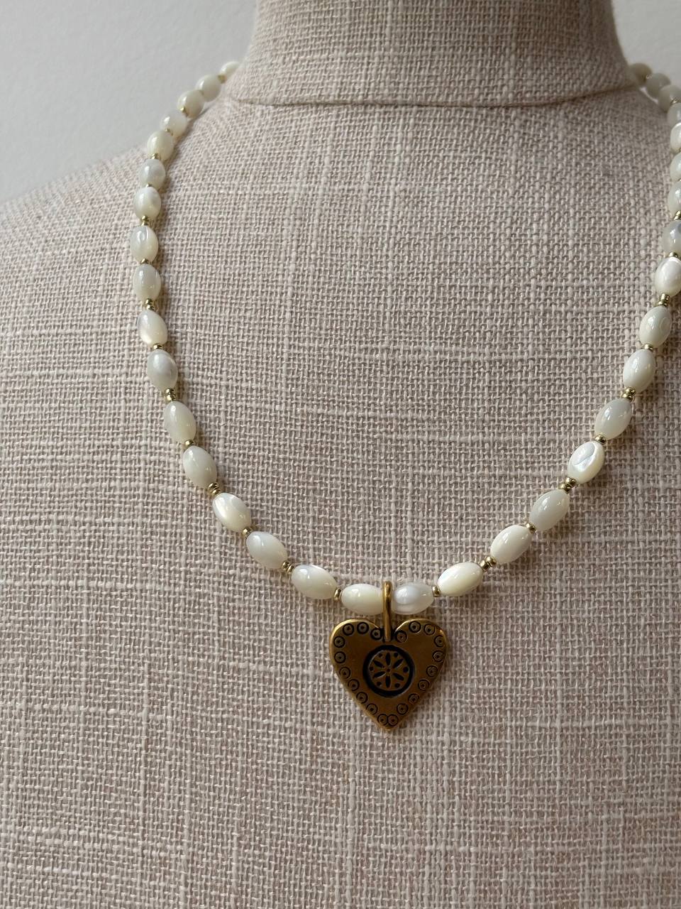 A necklace made of small white pearl-like beads with a gold-toned heart-shaped pendant featuring a sunburst design, displayed on a beige fabric mannequin bust.