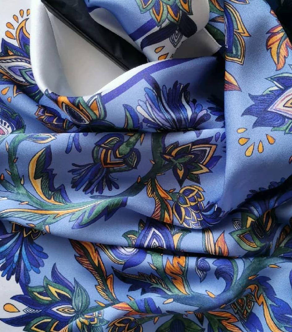 A close-up of a light blue patterned scarf or fabric with an intricate floral and geometric print in navy, green, orange, and yellow, draped to show its texture and design.