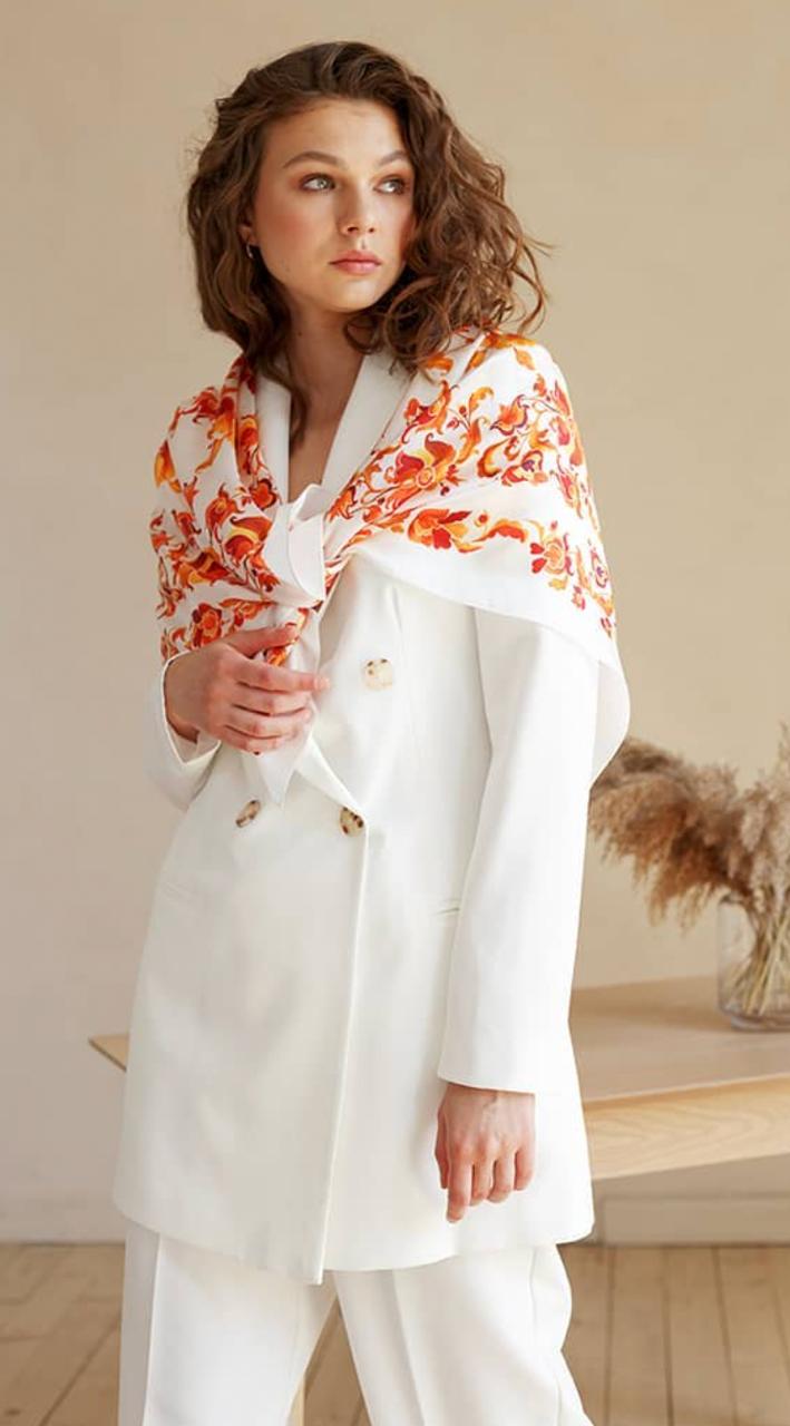white scarf featuring a vibrant orange and red floral pattern, posed in a warm, neutral-toned indoor setting.