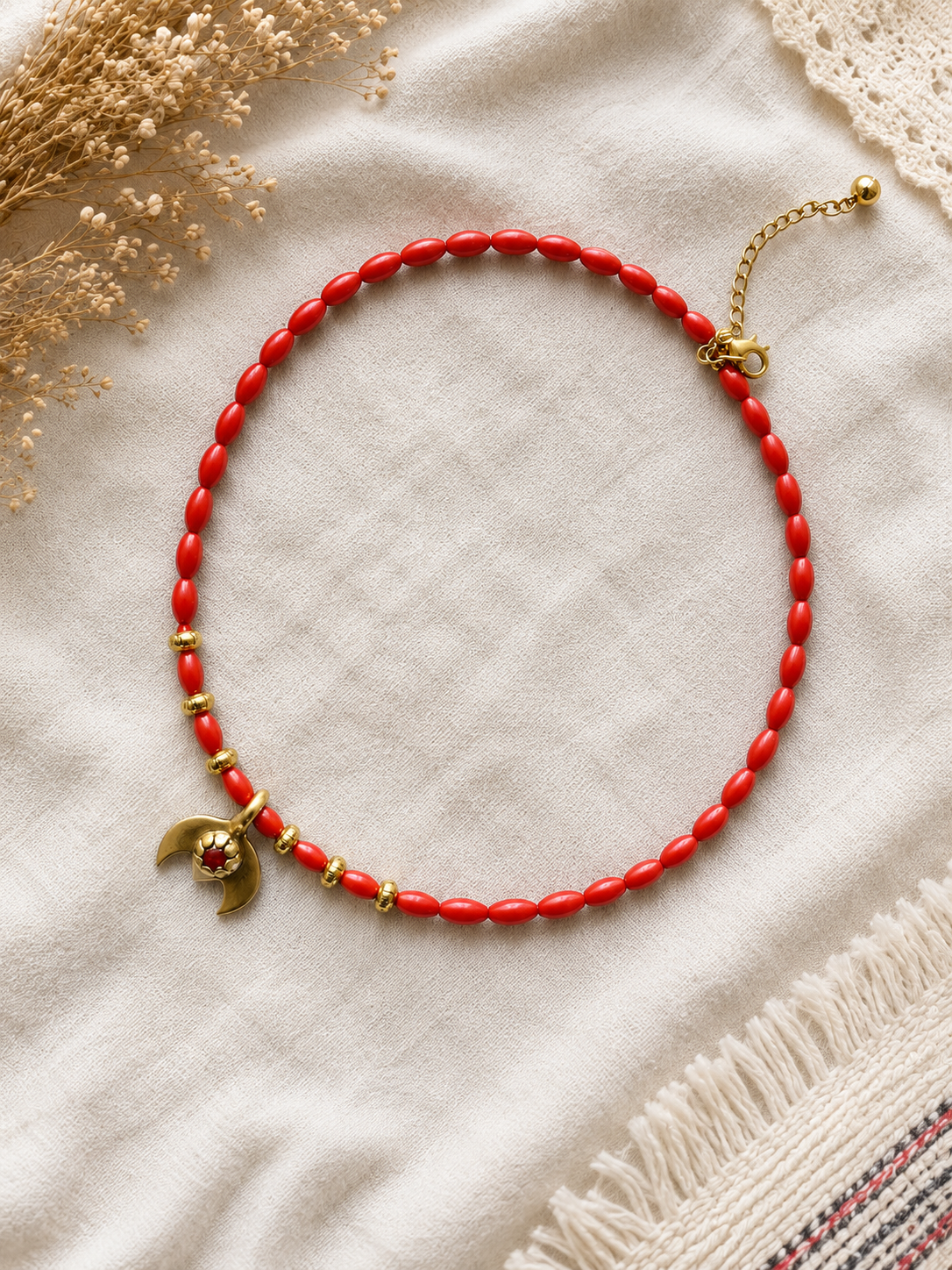 A red beaded necklace with gold-toned accents, including a crescent moon charm with a small red heart, displayed on a textured cream fabric with dried flowers and a lace trim.