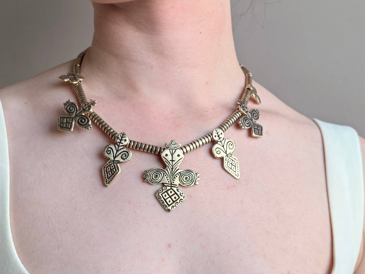 A close-up of a person wearing a chunky, ornate bronze-toned statement necklace with multiple geometric and tribal-inspired charms, paired with a white tank top.