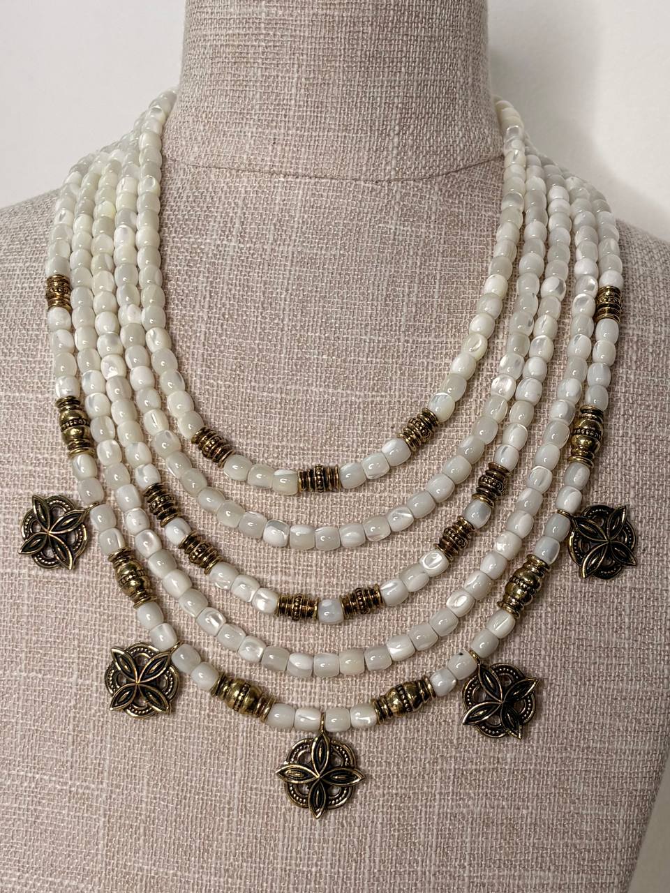 A multi-strand statement necklace displayed on a beige linen mannequin bust. The necklace features white mother-of-pearl beads, antique bronze cylindrical spacer beads, and antique bronze flower-shaped charms.
