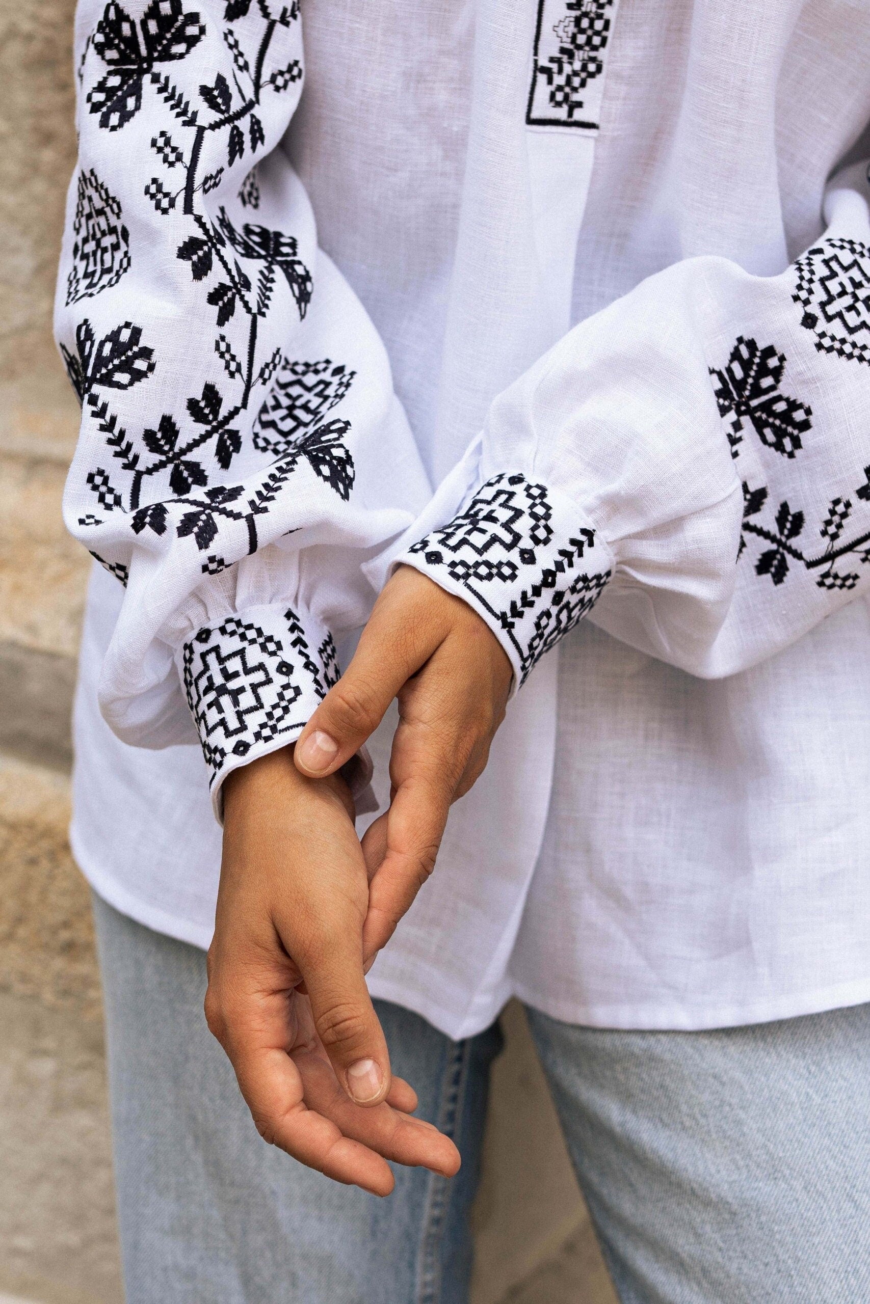 A close-up of a white linen blouse with black embroidered vyshyvanka patterns, sleeves, and a person's hands.
