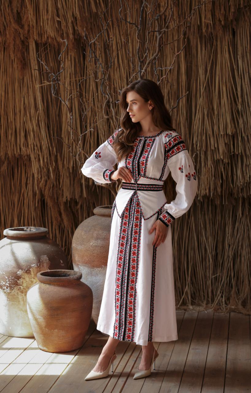 Woman in a white dress with red embroidery standing in front of a thatched wall.