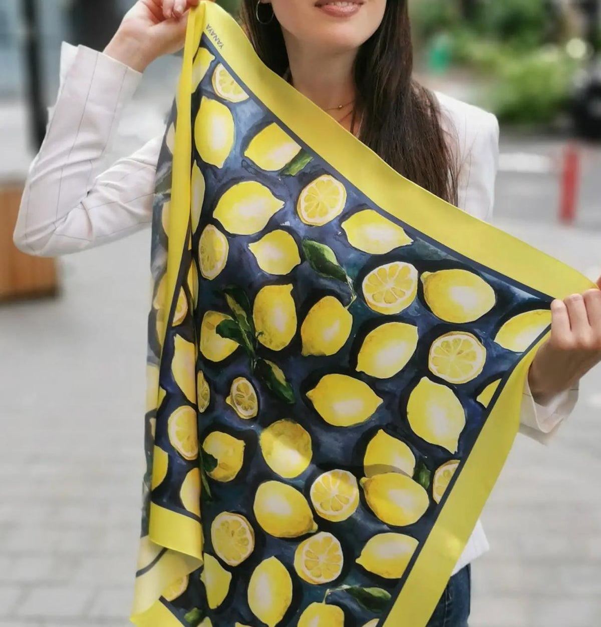 A woman holds up a square scarf featuring a vibrant yellow lemon pattern on a dark navy blue background, with a solid yellow border. She is wearing a white long-sleeve top and holding the scarf in an outdoor setting with a blurred background.