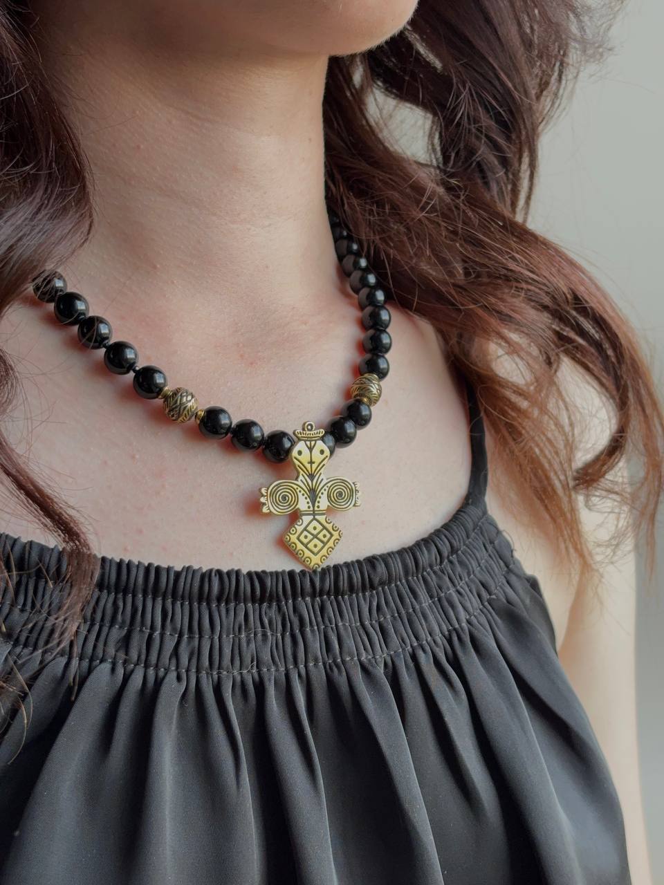 A close-up of a woman wearing a black beaded necklace with a gold-toned ornate pendant, paired with a dark gray sleeveless top.