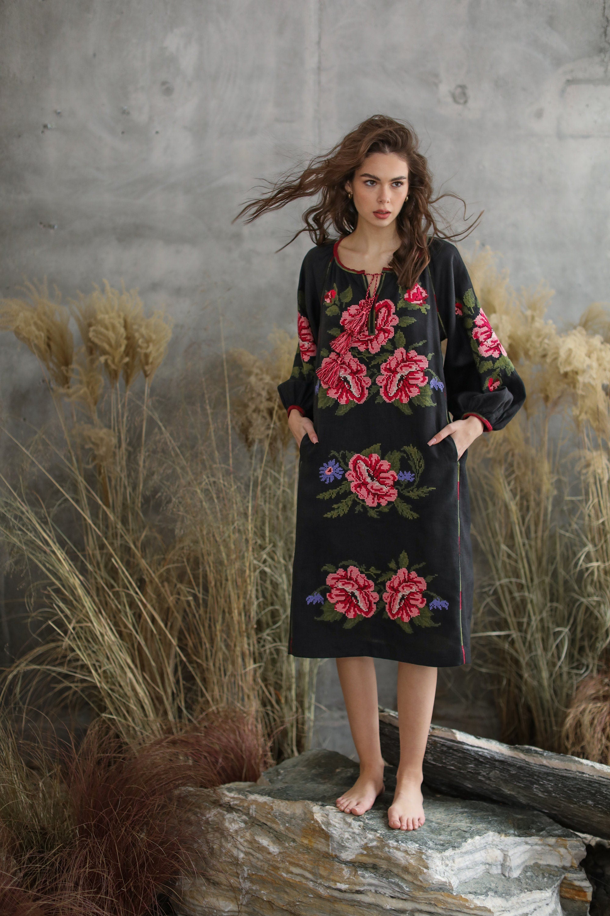 A woman standing on a rock, wearing a black floral embroidered dress with a bohemian ethnic style.