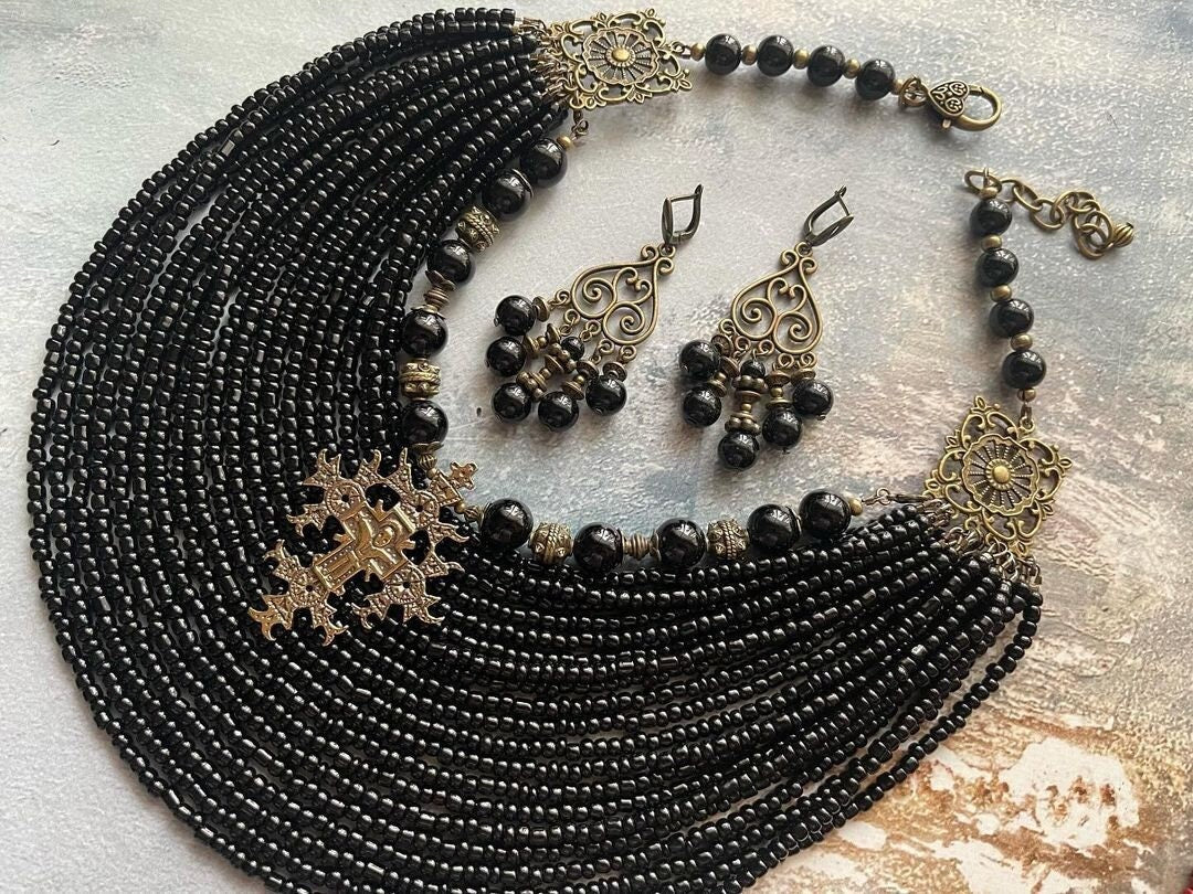 A multi-row black necklace made of glass beads with brass charms and metal hardware, accompanied by a pair of matching earrings.