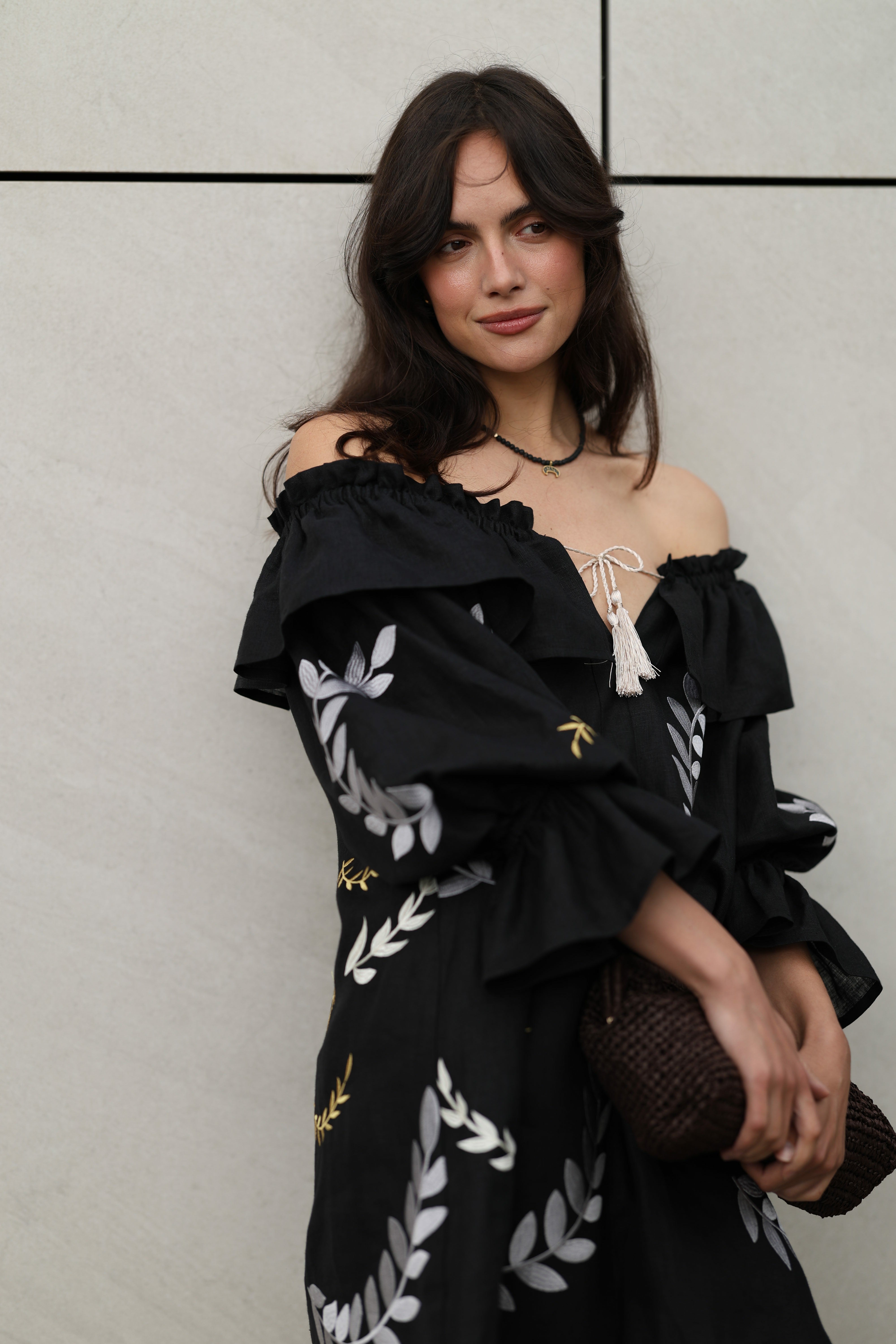Woman wearing a black off-shoulder dress with white floral patterns against a light gray wall.