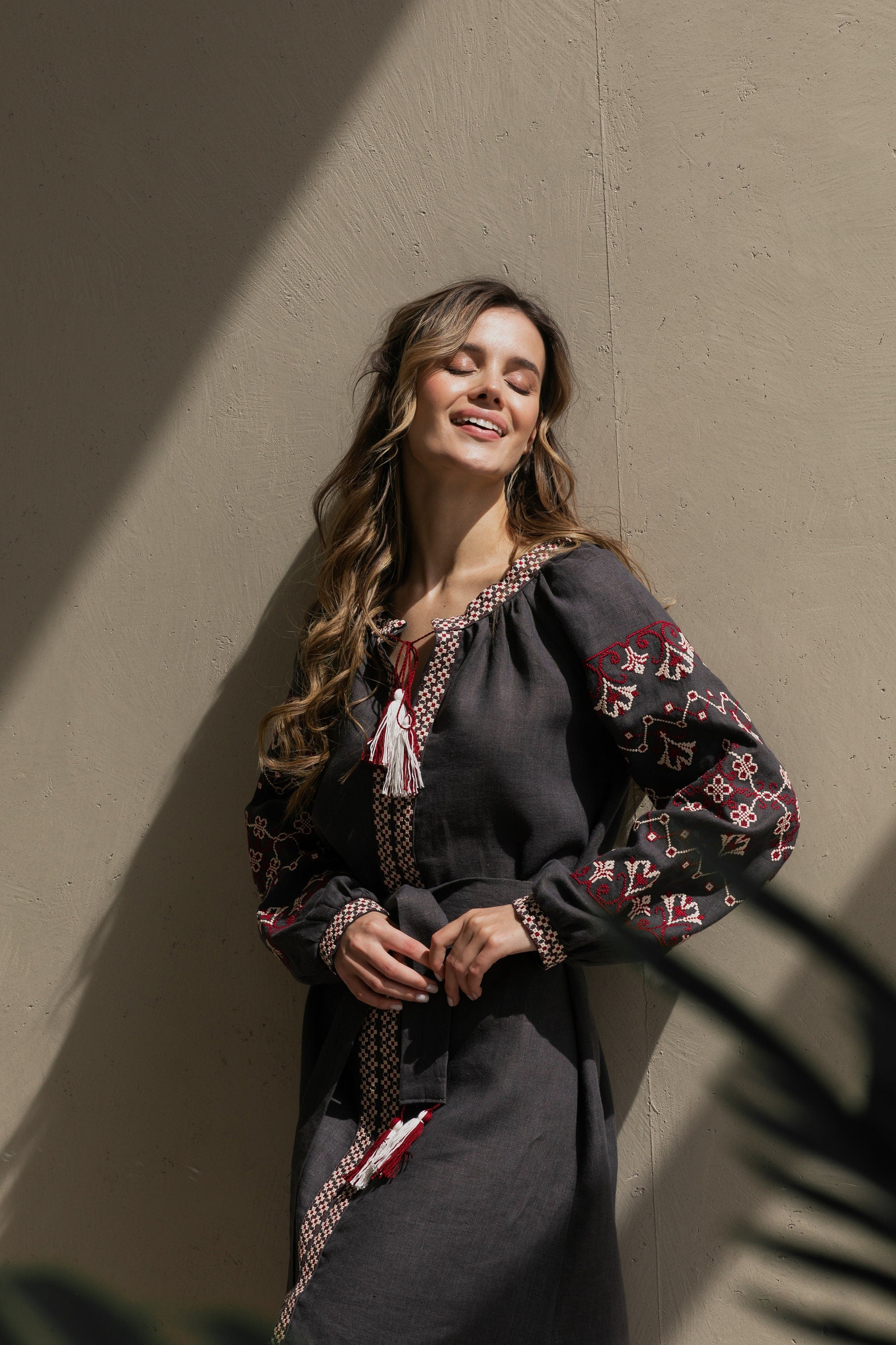 A woman standing in a shadowed space wearing a gray linen dress with Ukrainian embroidered vyshyvanka patterns in a boho folk style.