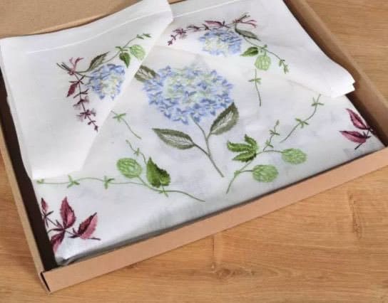 A linen tablecloth with embroidered hydrangea flowers displayed in an open box on a wooden surface.