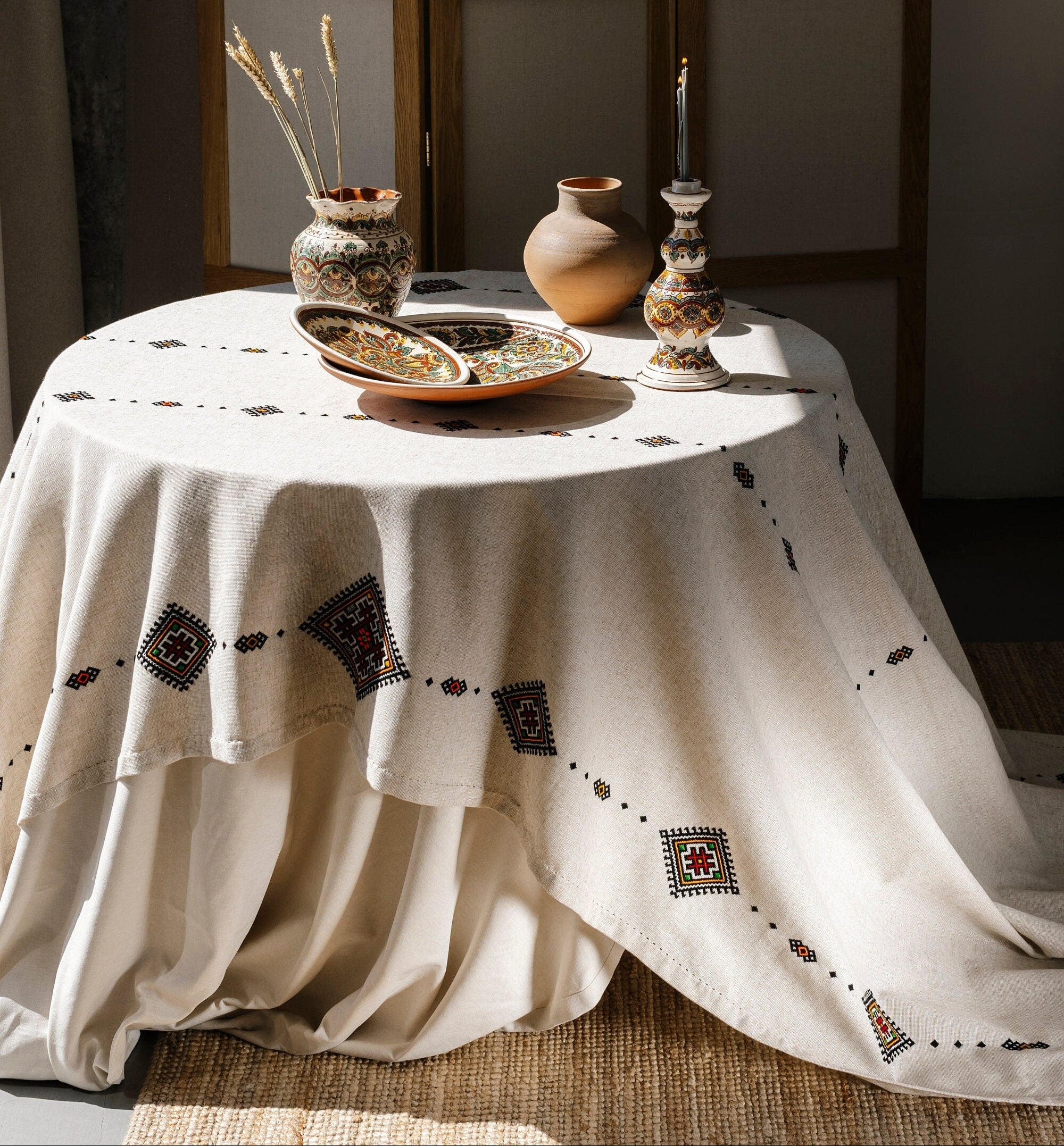 Handmade beige embroidered tablecloth with geometric patterns displayed on a table, complemented by decorative vases and candles.