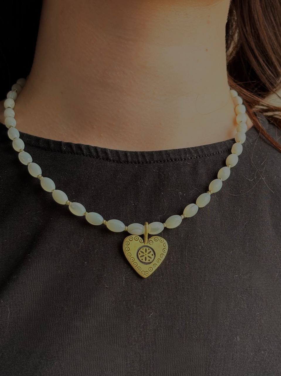 A close-up of a person's neck and chest wearing a beaded necklace with a gold heart-shaped pendant featuring a wheel design.