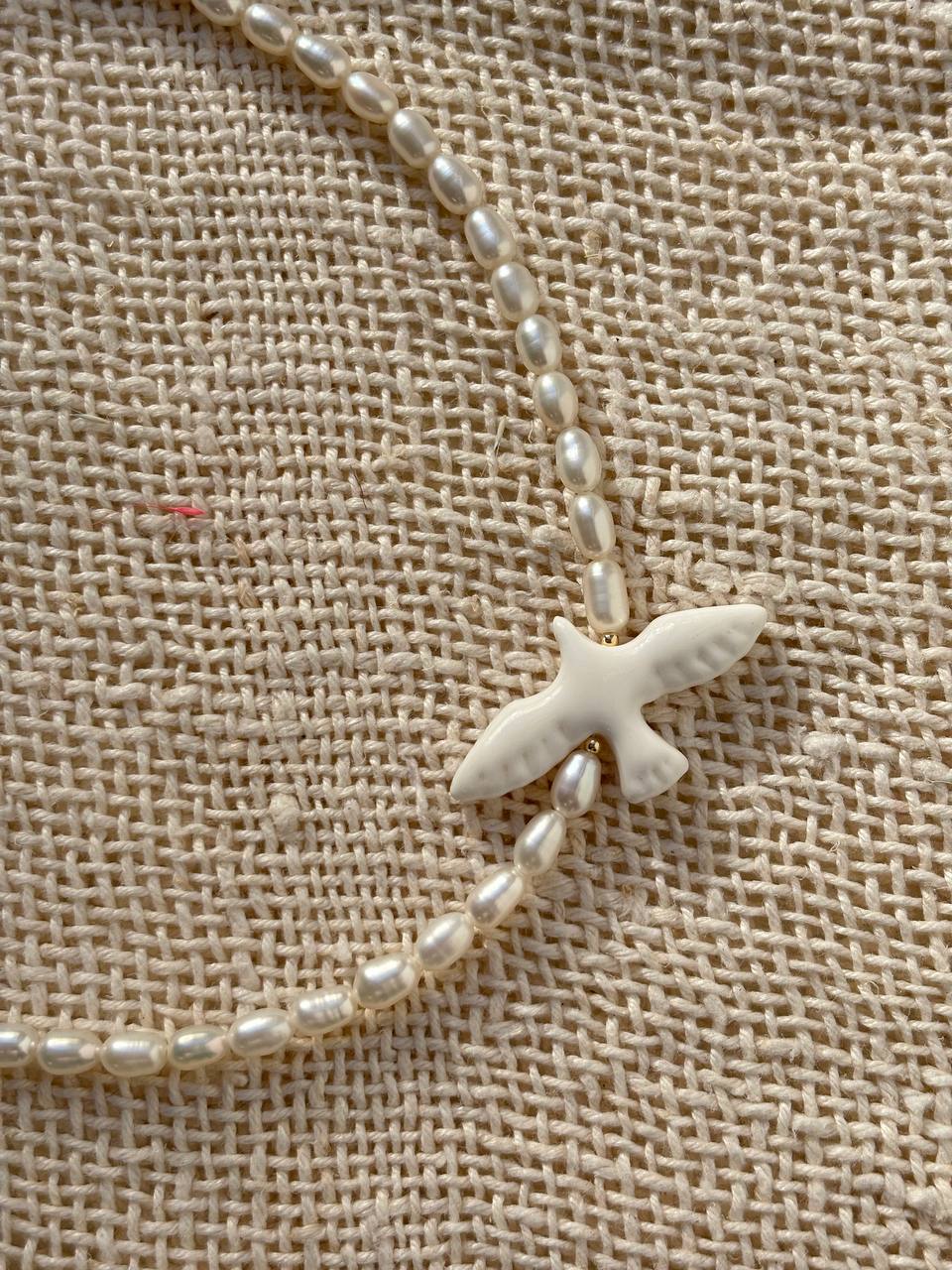 A close-up of a necklace with small white pearl-like beads and a white bird-shaped pendant, laid on a beige woven fabric background.