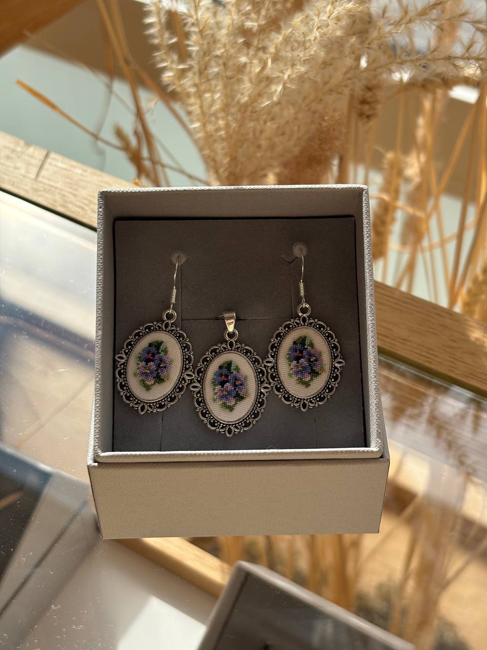 A pair of dangling earrings displayed in an open jewelry box. Each earring has an oval silver-toned filigree frame with a white center featuring purple grapes and green leaves, hanging from silver-toned hooks.