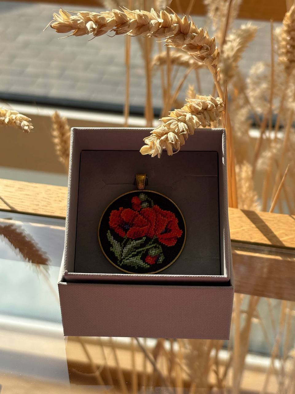 A round embroidered patch featuring red poppies with green leaves on a black background, presented inside an open gray gift box. The box is styled with dried wheat stalks against a warm, sunlit outdoor background.