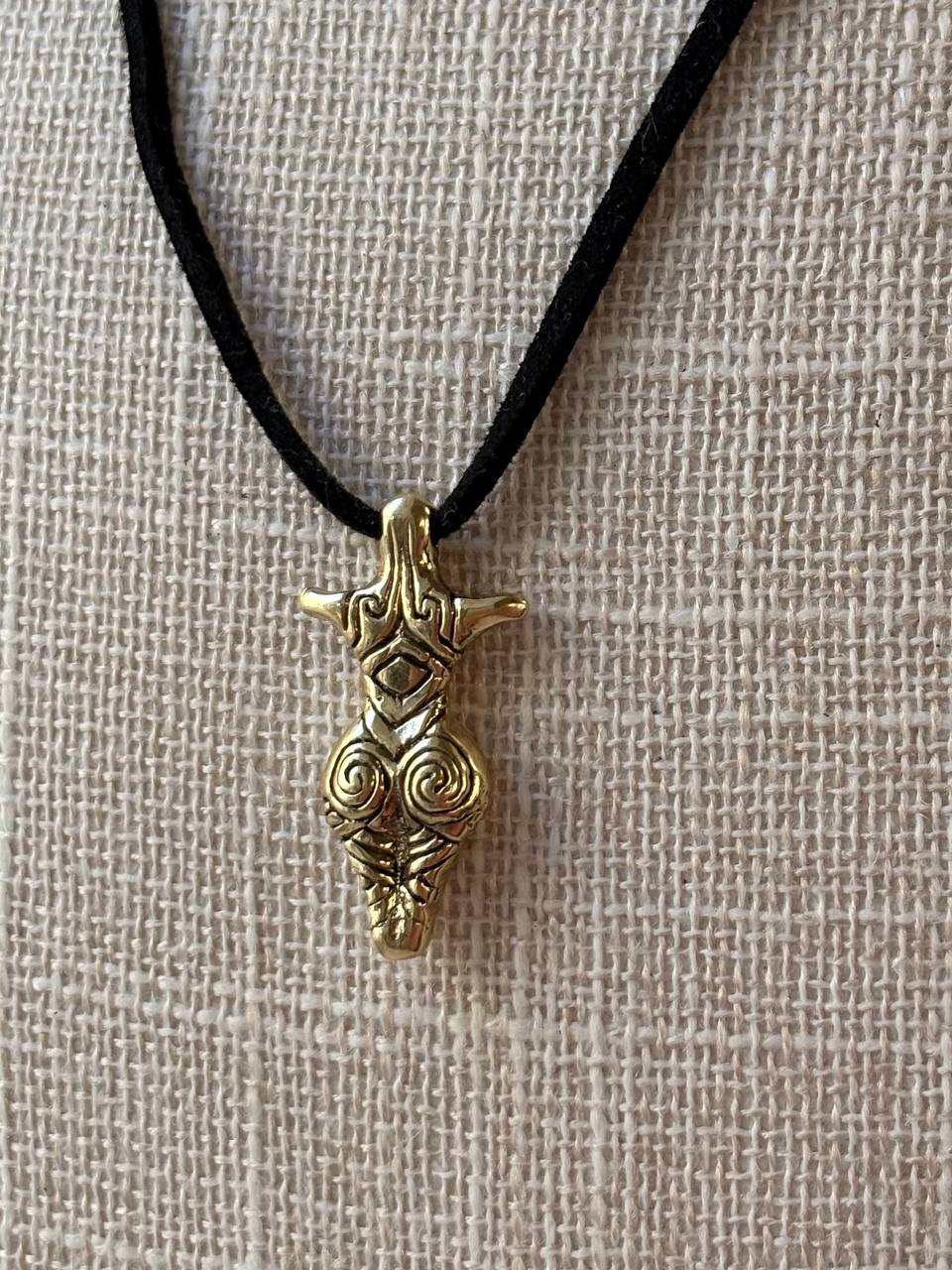 A gold-toned pendant with intricate Norse-style knotwork and a horned animal motif, hanging from a black cord necklace on a textured beige linen background.
