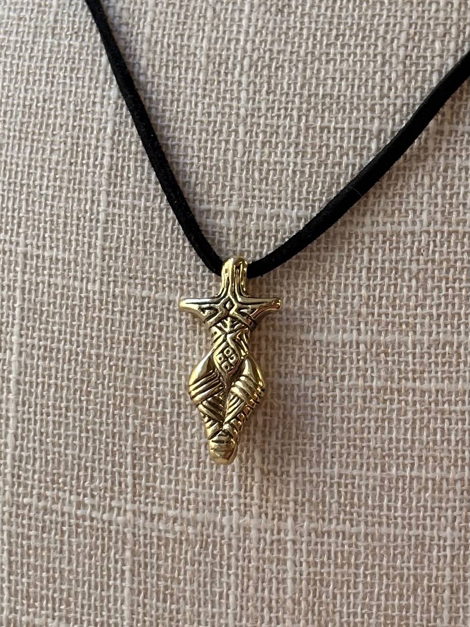 A gold-toned metal pendant with intricate carved details hangs from a black cord necklace, displayed against a textured beige linen background.
