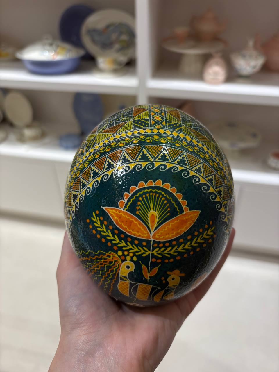 A hand holds a decorated Easter egg in a ceramic shop. The egg features a dark teal-green base with intricate hand-painted patterns in yellow, orange, and white, including traditional Ukrainian folk art motifs like a stylized sun, a peacock, and geometric borders.