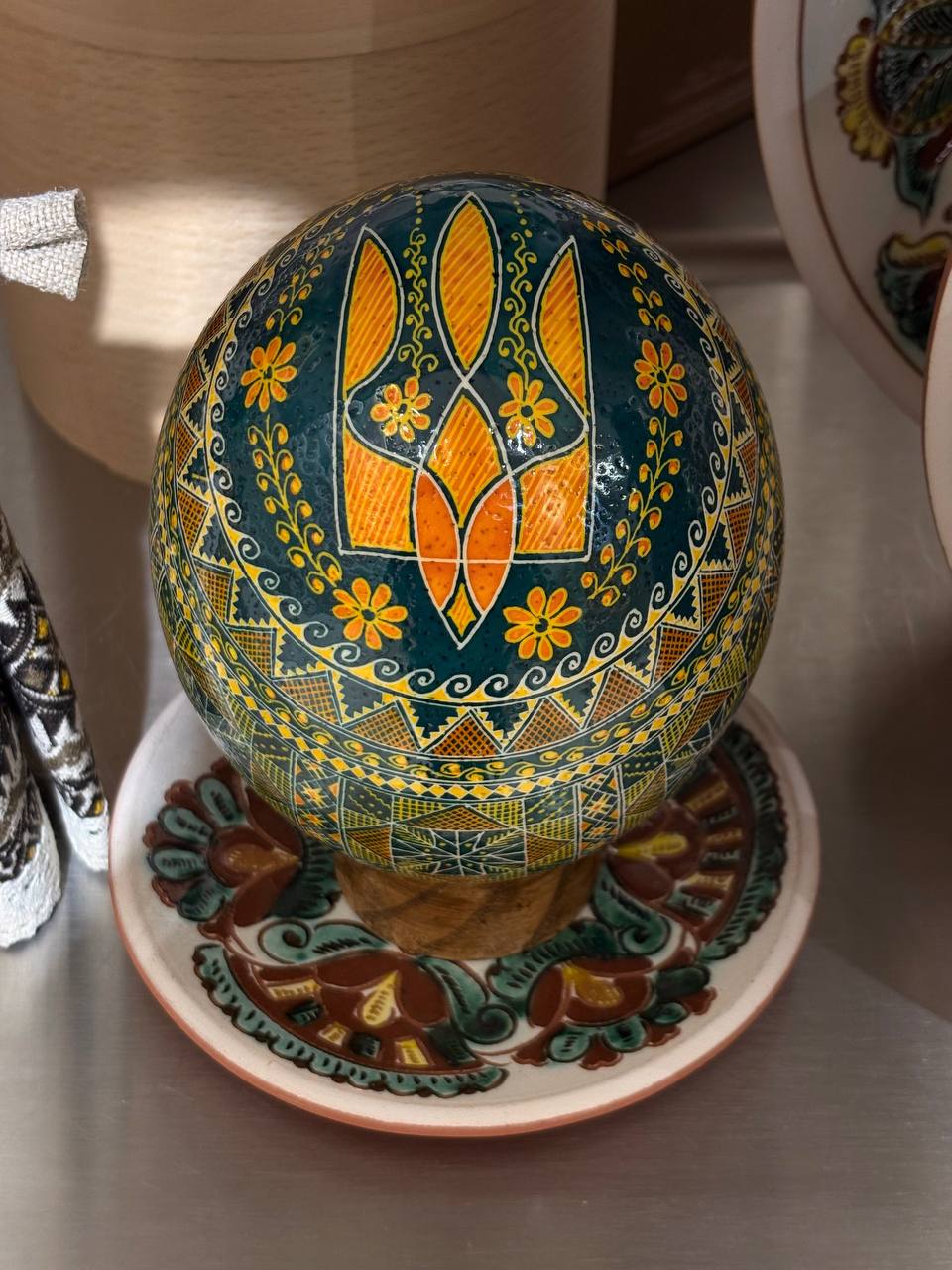 A hand-painted Ukrainian pysanka egg with a blue-green background, yellow floral motifs, and geometric patterns, displayed on a matching ceramic stand.