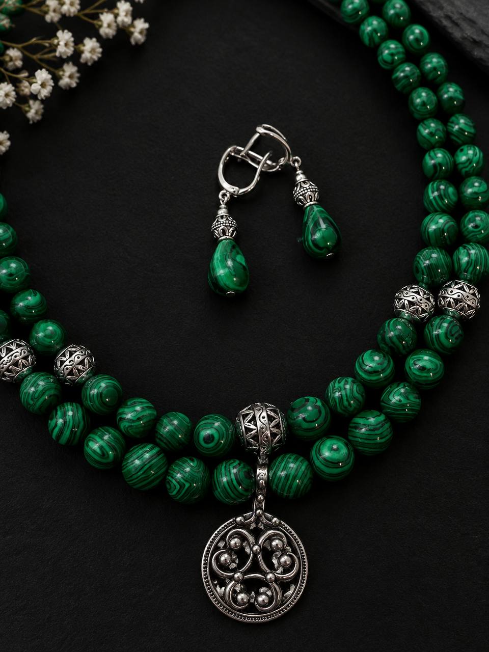 A green beaded necklace and matching drop earrings with ornate silver-toned metal details, displayed on a dark textured background with small white flowers in the corner.