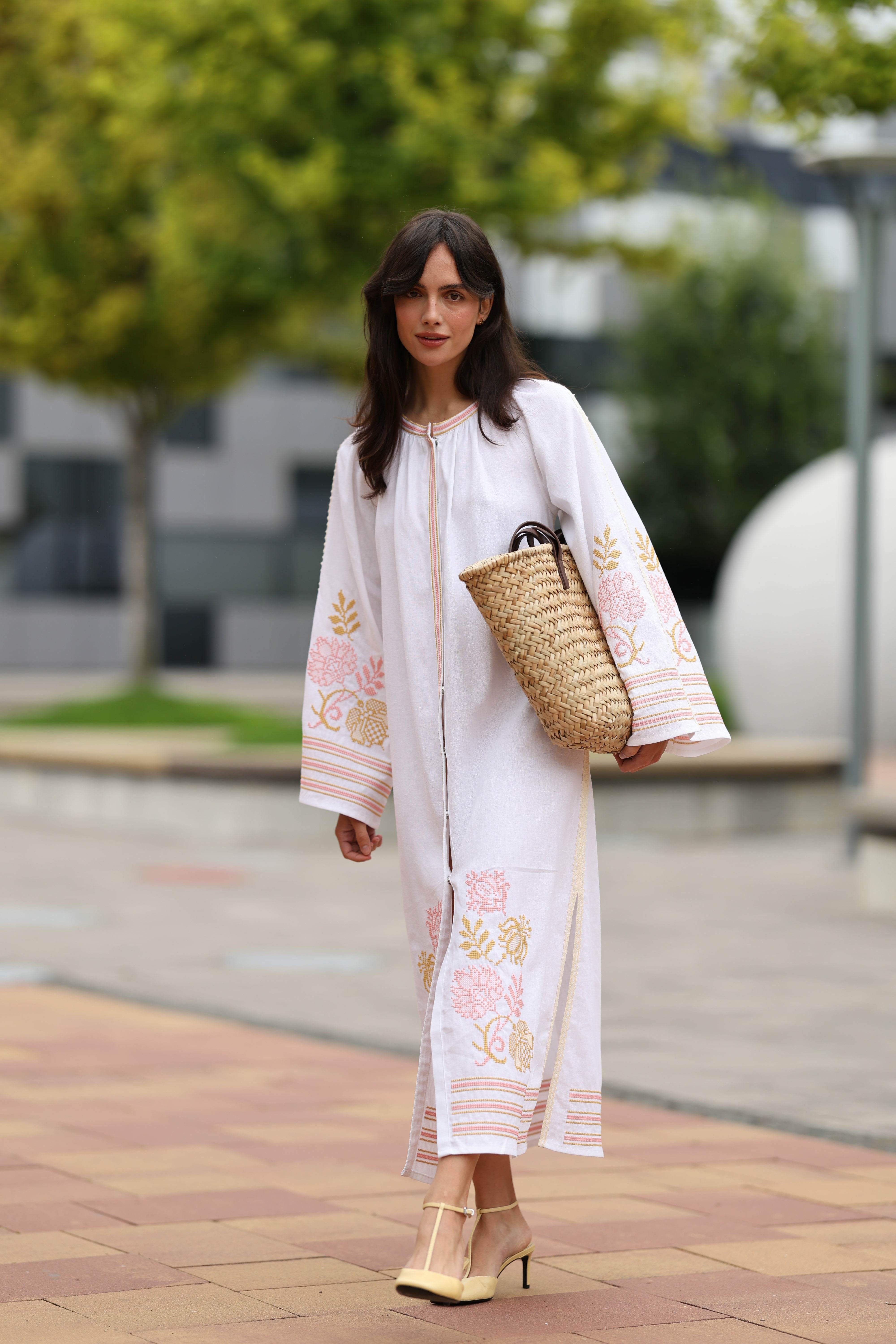 Woman in a white embroidered dress holding a straw bag on a city street.