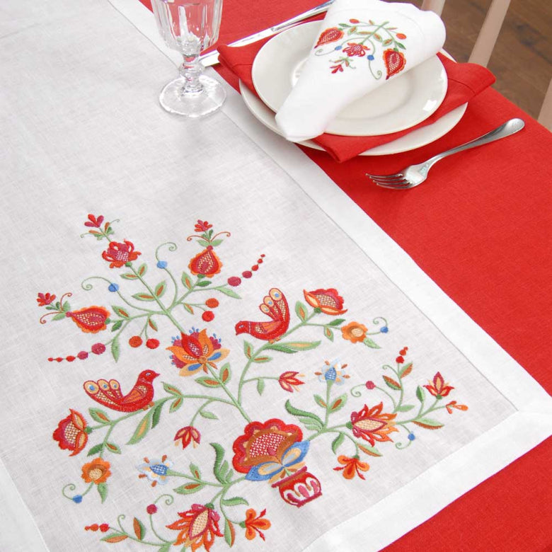 A white embroidered table runner with a colorful floral and bird pattern, styled on a red tablecloth with white plates, a folded napkin, and silverware.