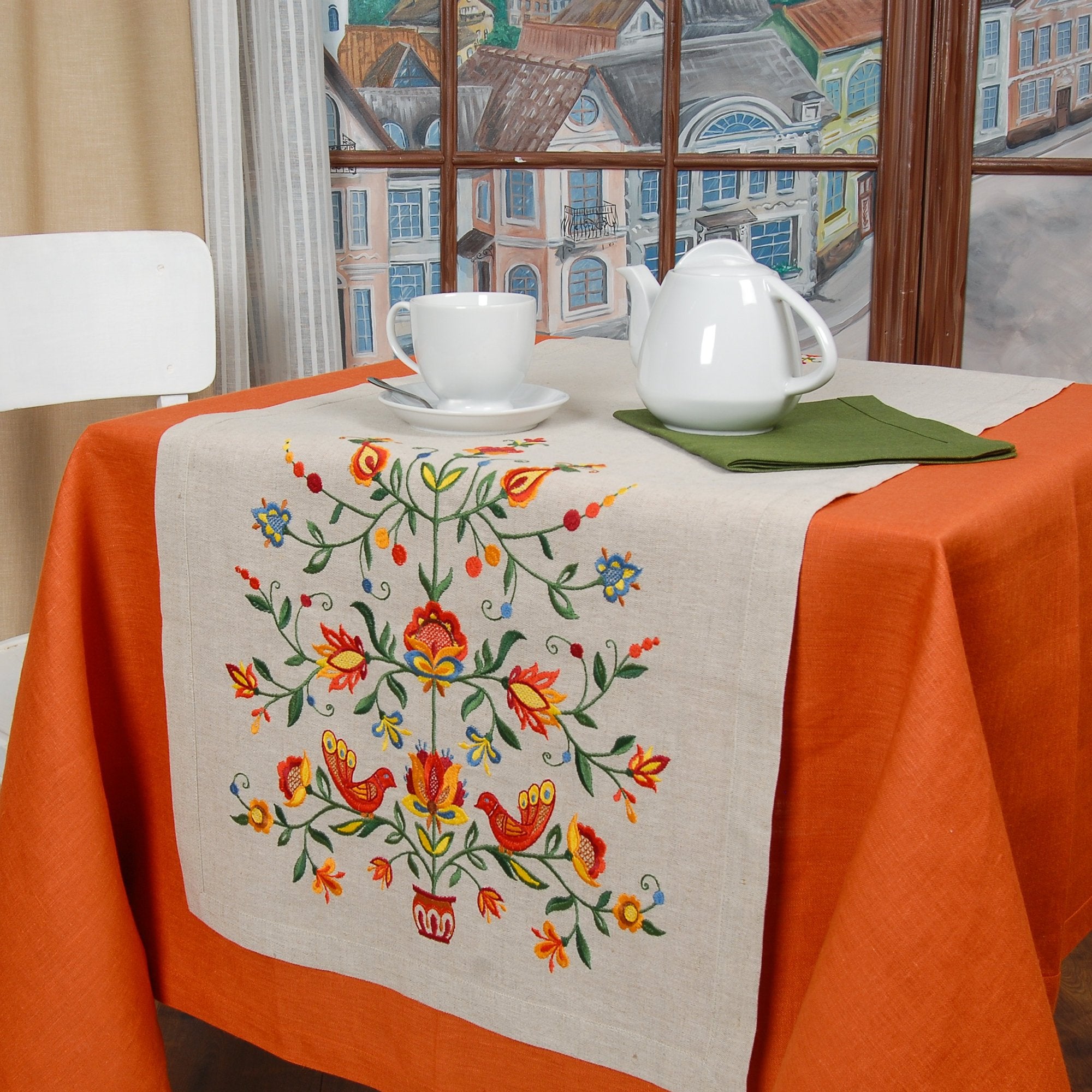 An embroidered tablecloth with a floral Tree of Life design displayed on a table with tea set and decorative items in the background.