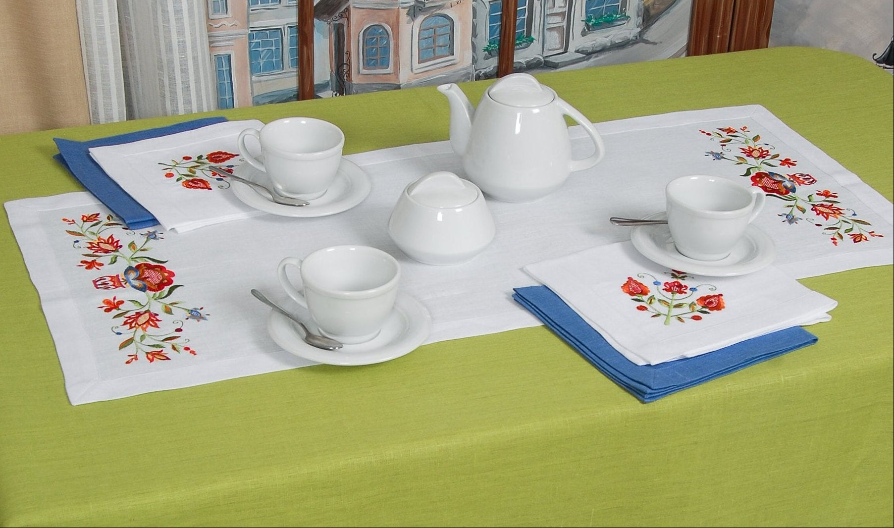 An embroidered tablecloth with a floral pattern displayed on a green table, setting includes dishes and cutlery.