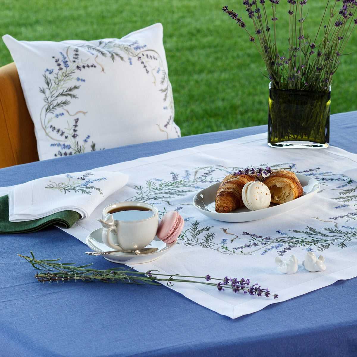 A linen tablecloth featuring a floral pattern in a blue and green color palette, displayed on a table with a cup of coffee and baked goods.
