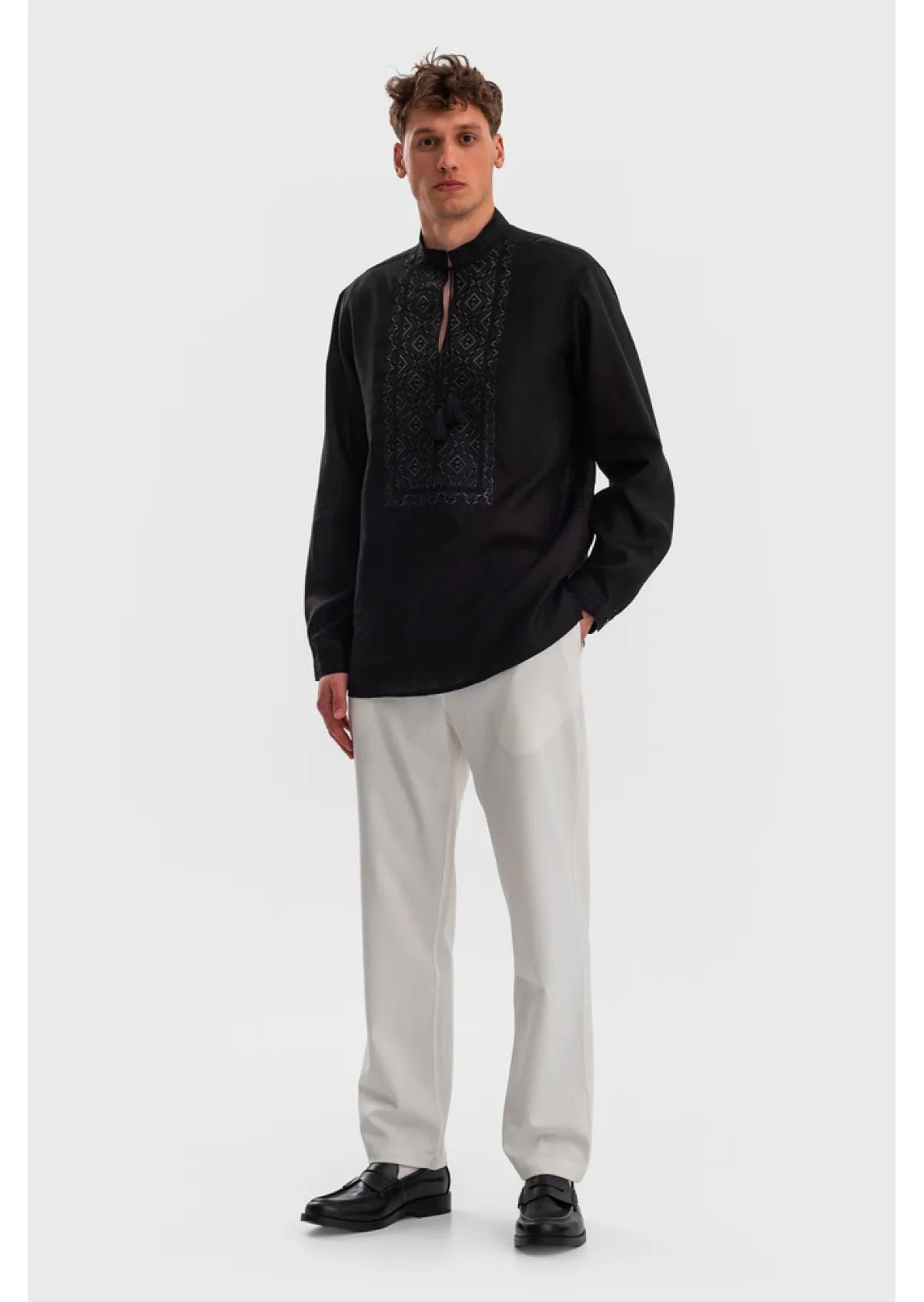 A man modeling a black long-sleeve kurta with dark embroidery on the chest, paired with off-white trousers and black loafers, against a light gray studio background.