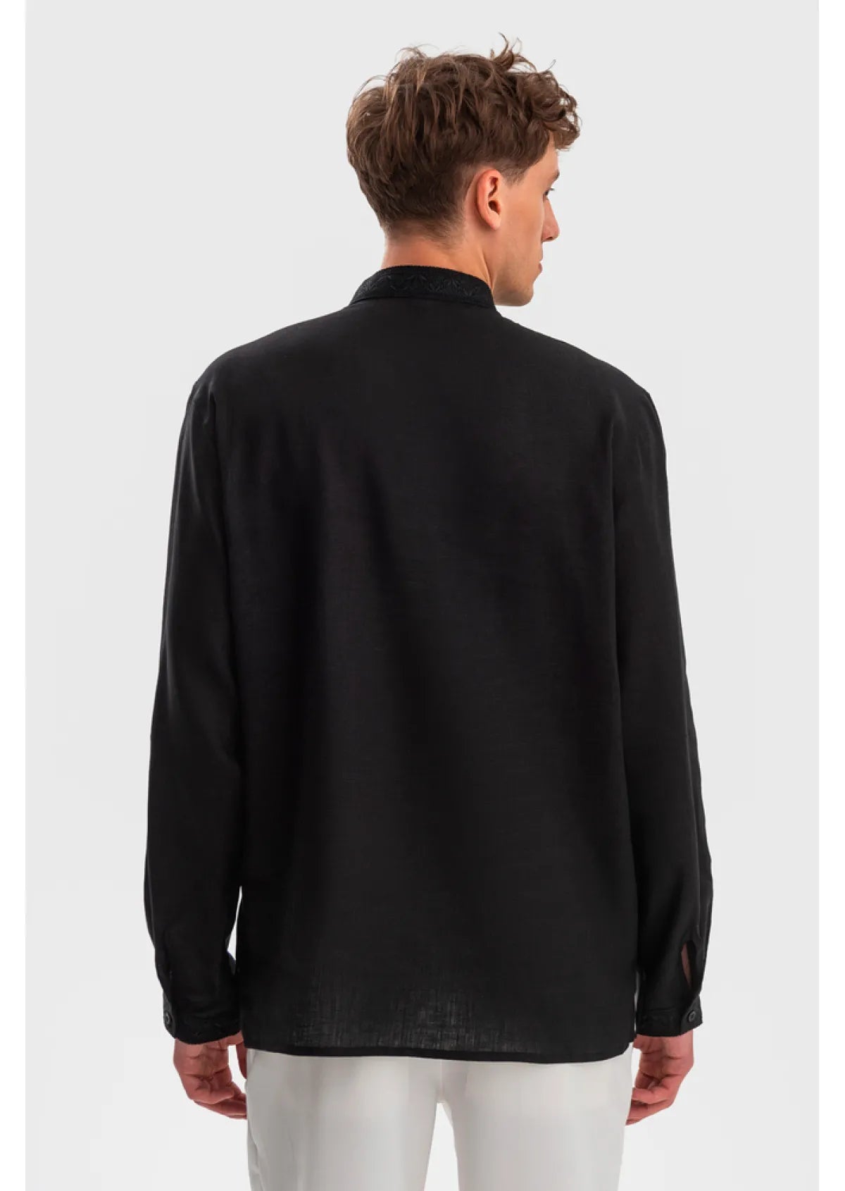 A man viewed from the back wearing a black long-sleeve shirt and white pants against a light gray studio background.