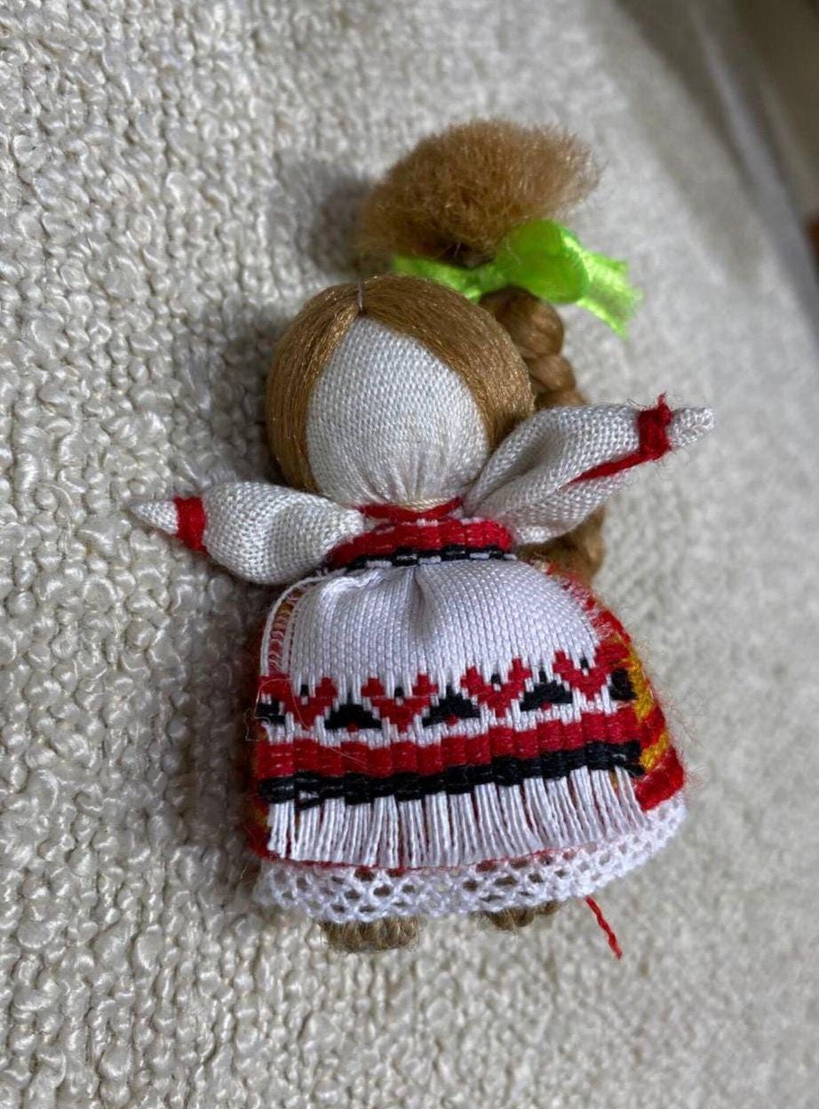 A small handmade doll with a predominantly white and red outfit, featuring embroidered details and a green ribbon, crafted from fabric.