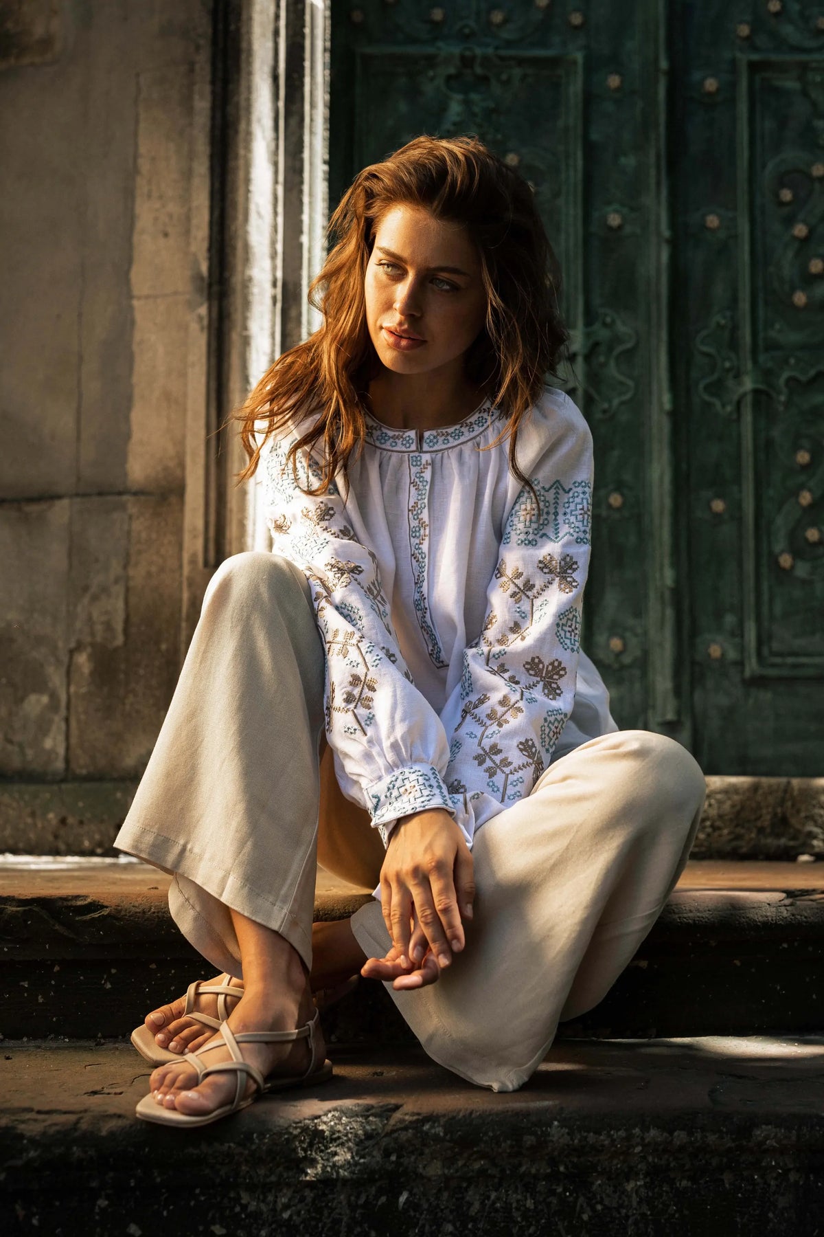 A woman sits on stone steps in front of a dark green wooden door, wearing a white embroidered long-sleeve blouse and light beige wide-leg pants with beige sandals.