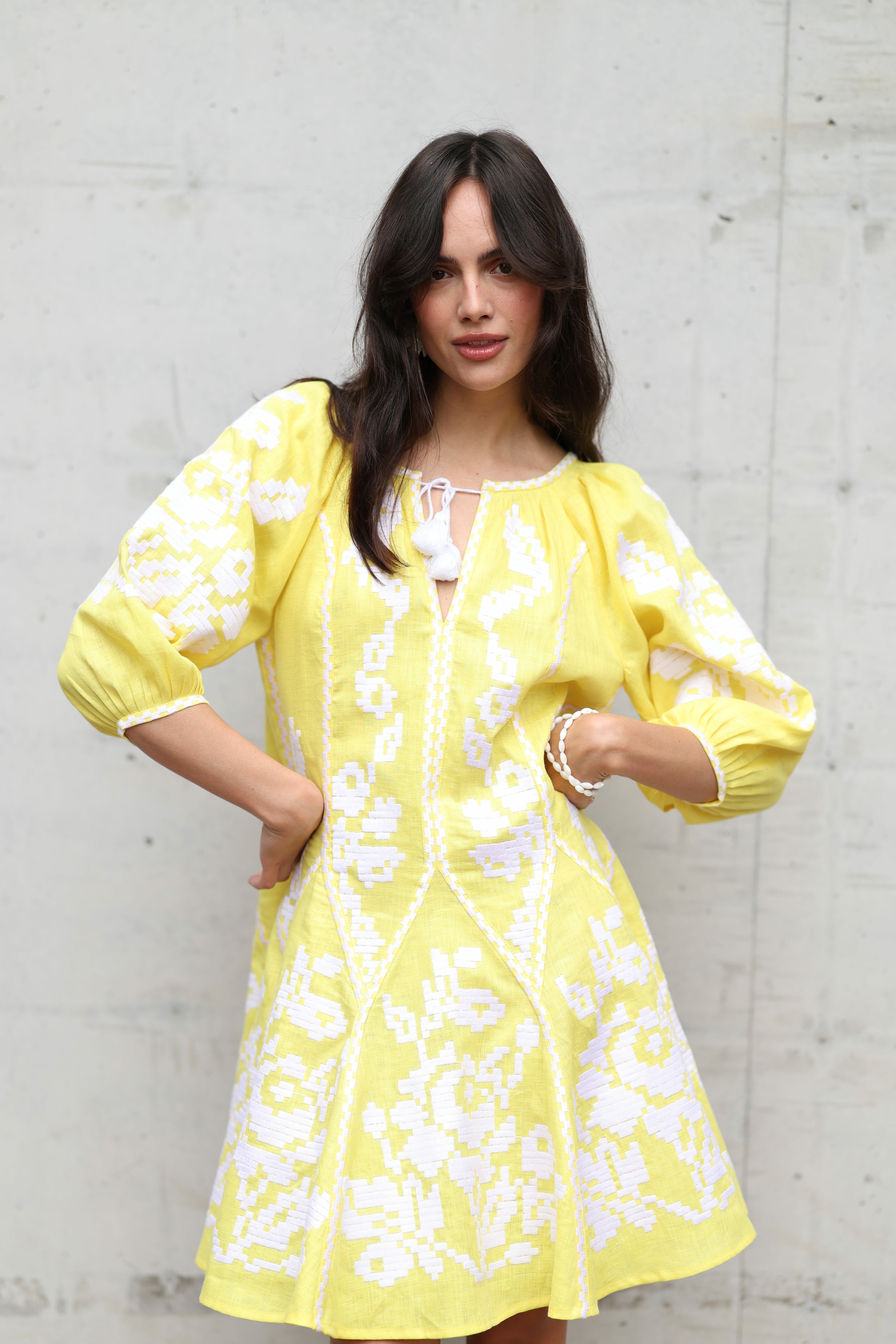 Woman wearing a yellow dress with white floral patterns against a gray concrete wall.