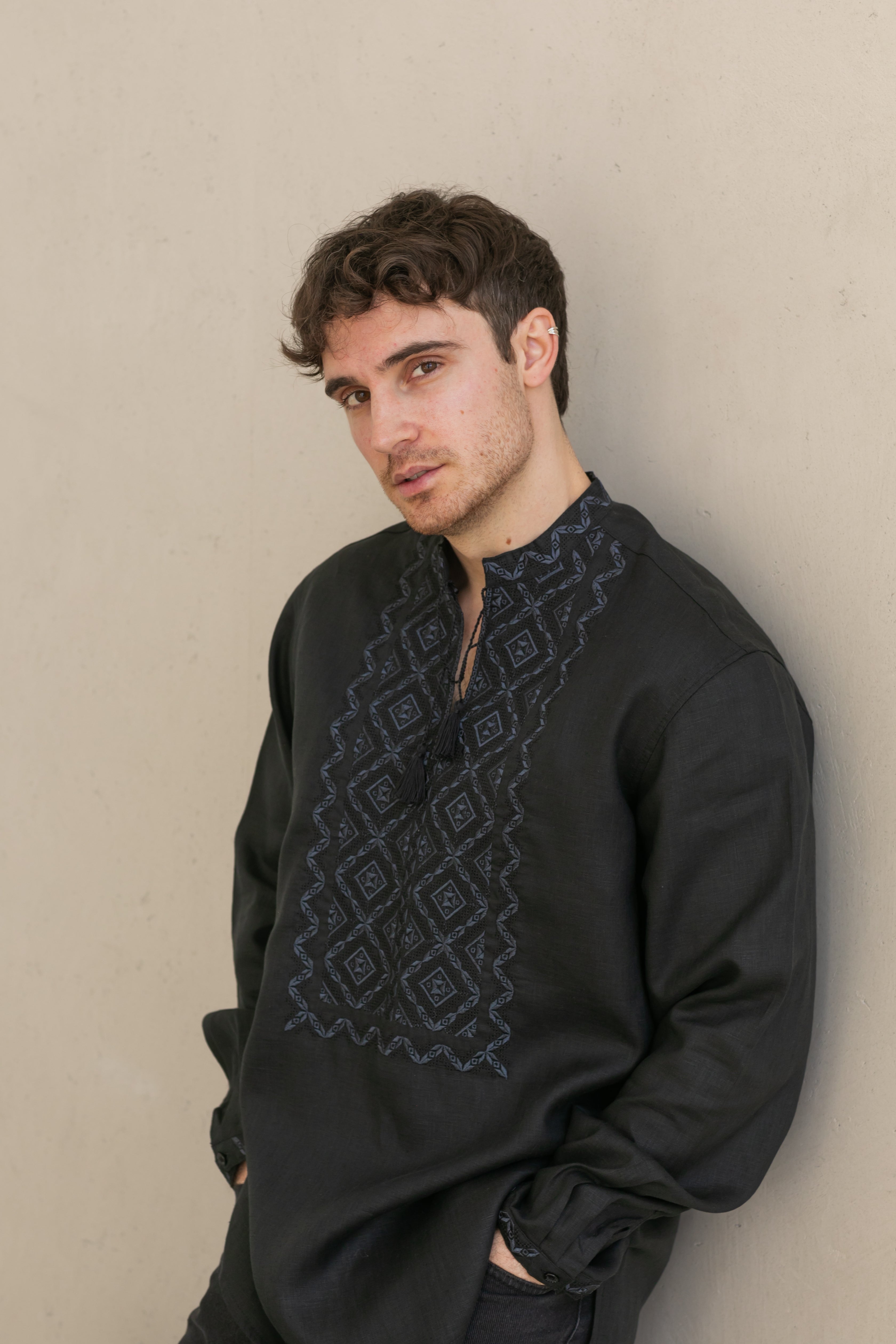 Man wearing a black embroidered shirt against a beige wall