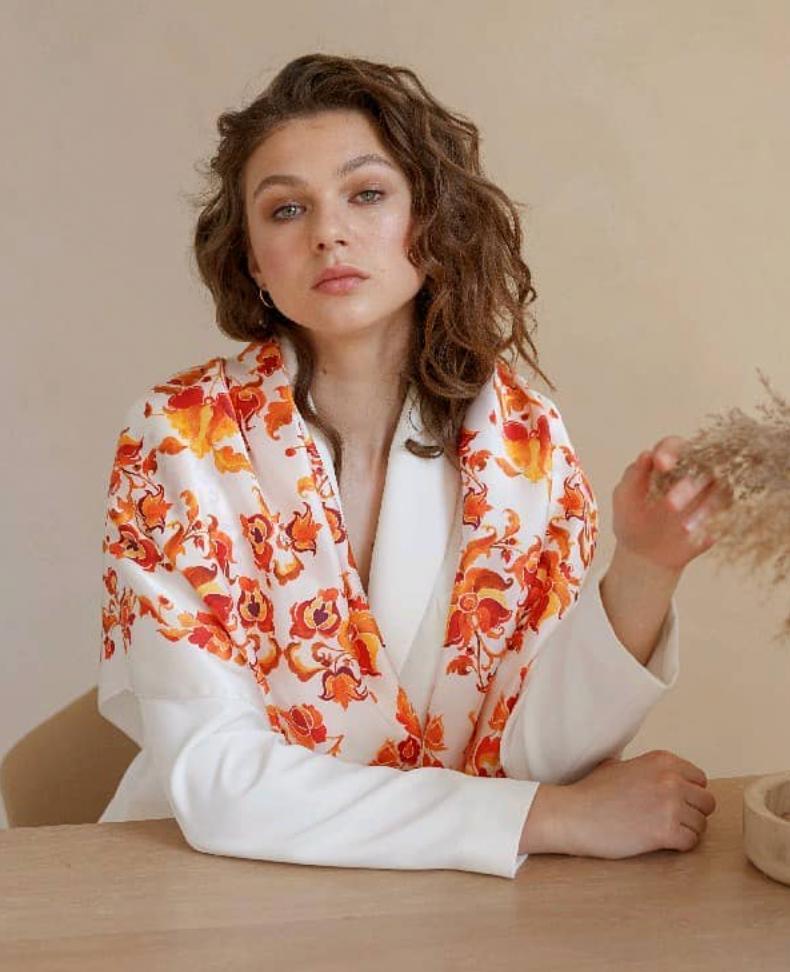 A woman with wavy brown hair sits at a light wood table wearing a white long-sleeve top and a white shawl draped over her shoulders, decorated with a vibrant orange and red floral pattern. She is holding a small bunch of dried grass against a neutral beige background.