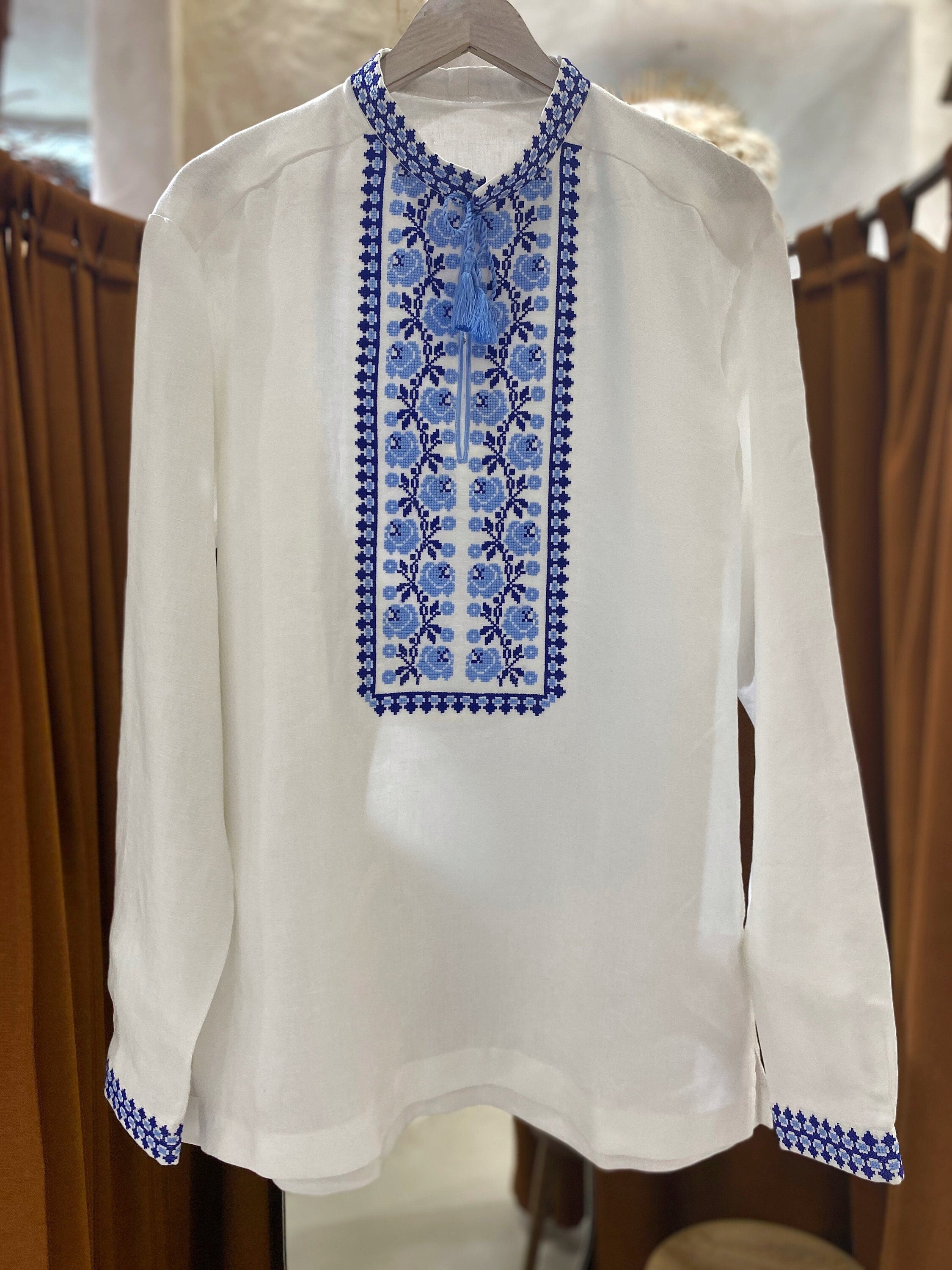 Men's white linen vyshyvanka shirt with blue embroidery, displayed against a hanging display setup.