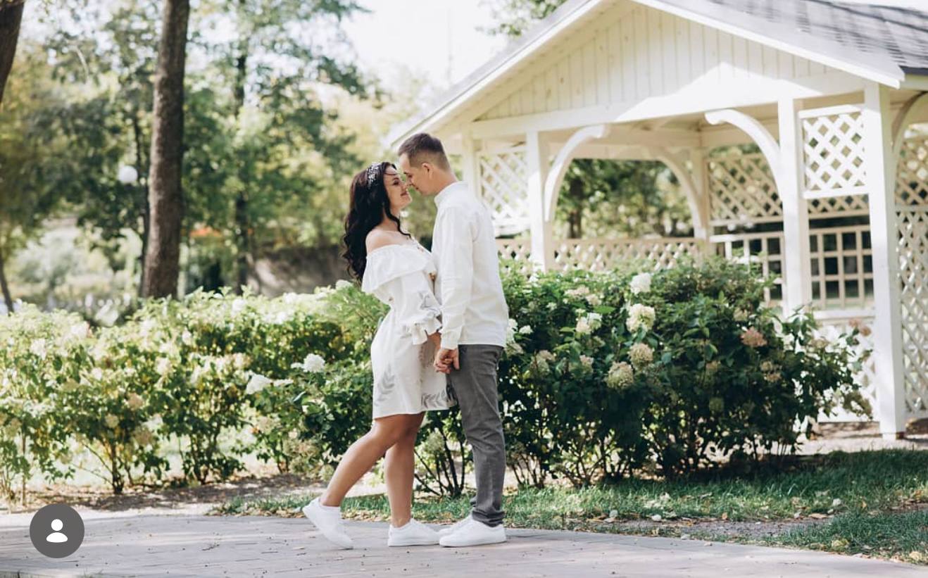 A couple embracing and kissing in a park setting with a white wooden gazebo and green bushes in the background. The woman is wearing a white off-the-shoulder dress and white sneakers, and the man is wearing a white shirt, gray pants, and white sneakers.
