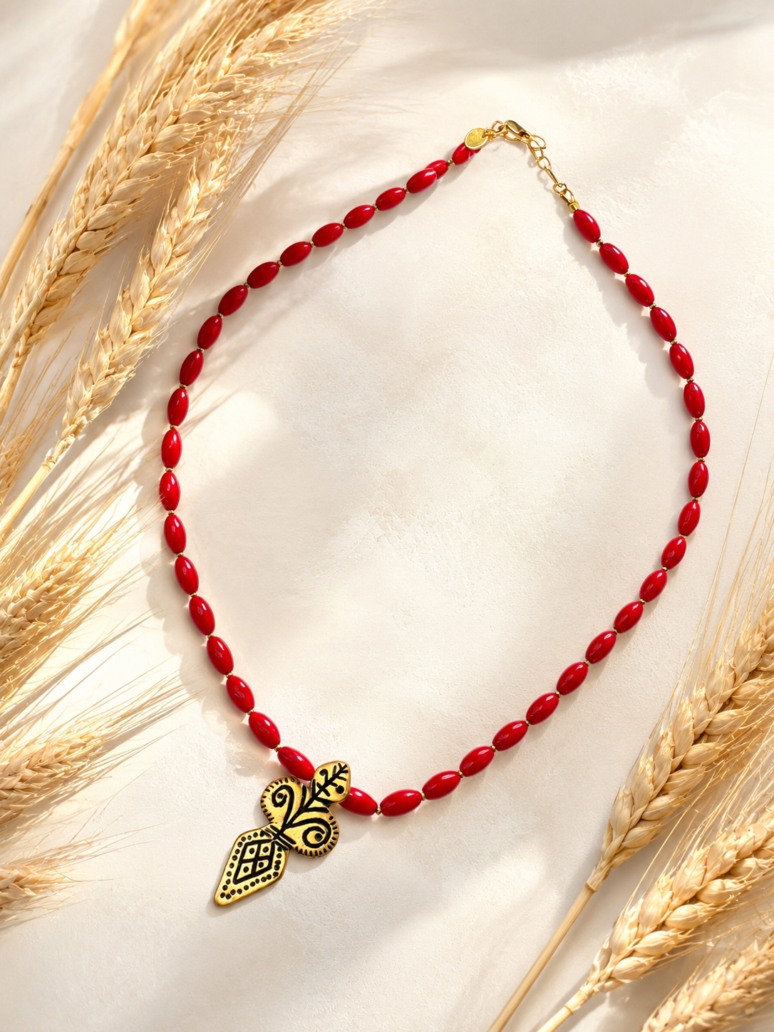 A red beaded necklace with a gold-toned ornate pendant, styled on a light cream fabric with dried wheat stalks.