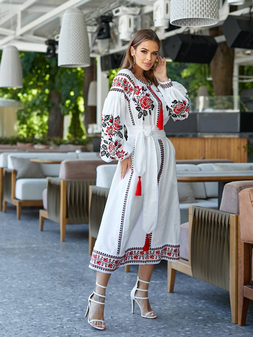 Woman in a white dress with red and black floral embroidery standing in an outdoor setting.