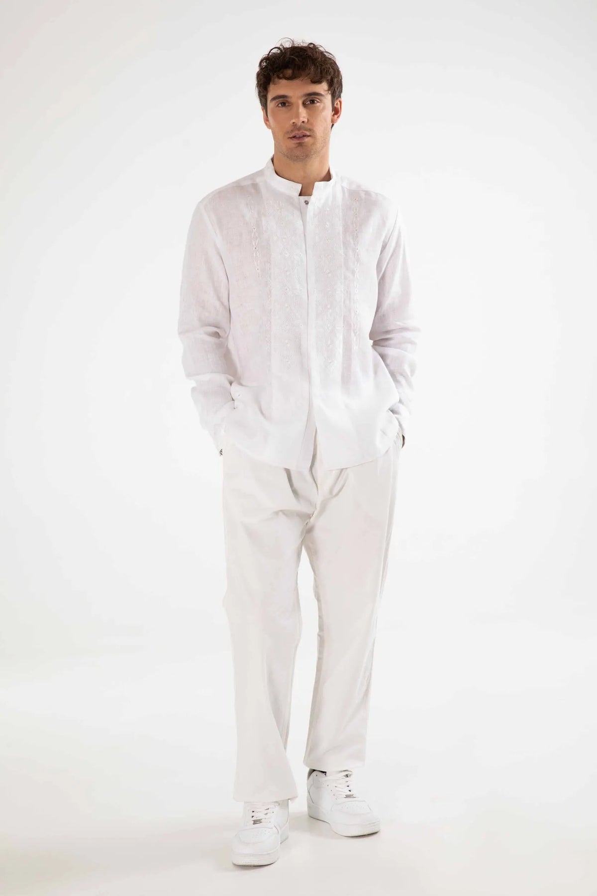 A man modeling a white long-sleeve linen shirt with a mandarin collar, paired with white trousers and white sneakers, against a plain white studio background.