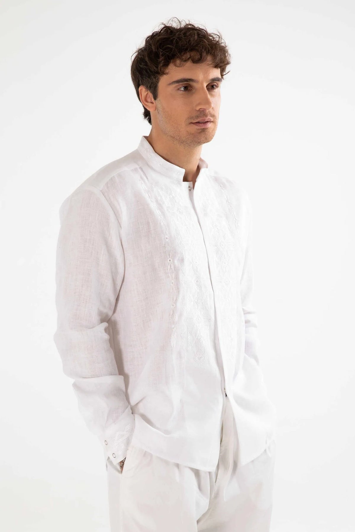 A man wearing a white long-sleeve linen shirt with a mandarin collar and white trousers, standing against a plain white background.