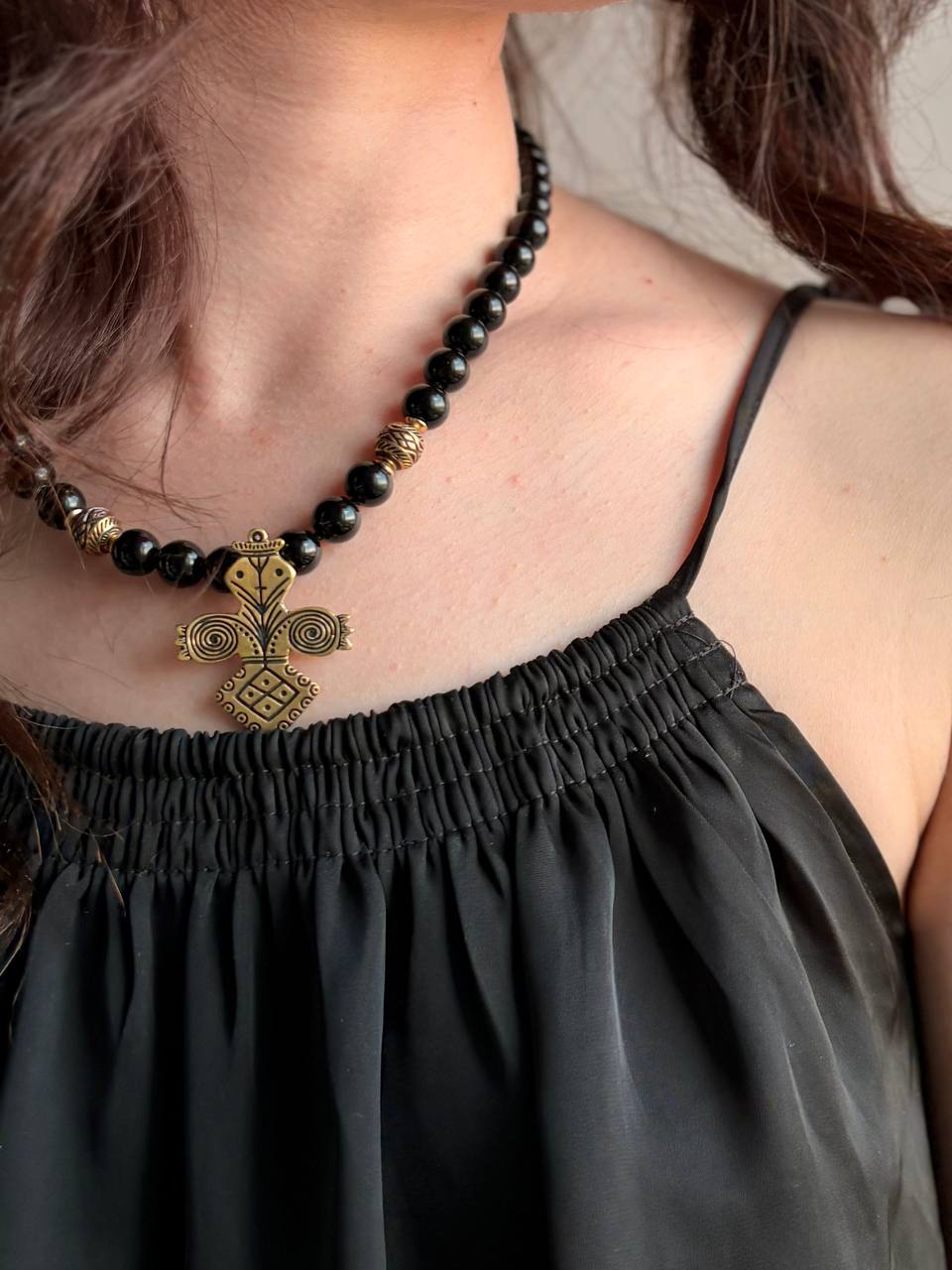 A close-up of a person wearing a black beaded necklace with a gold-toned ornate pendant, paired with a black spaghetti