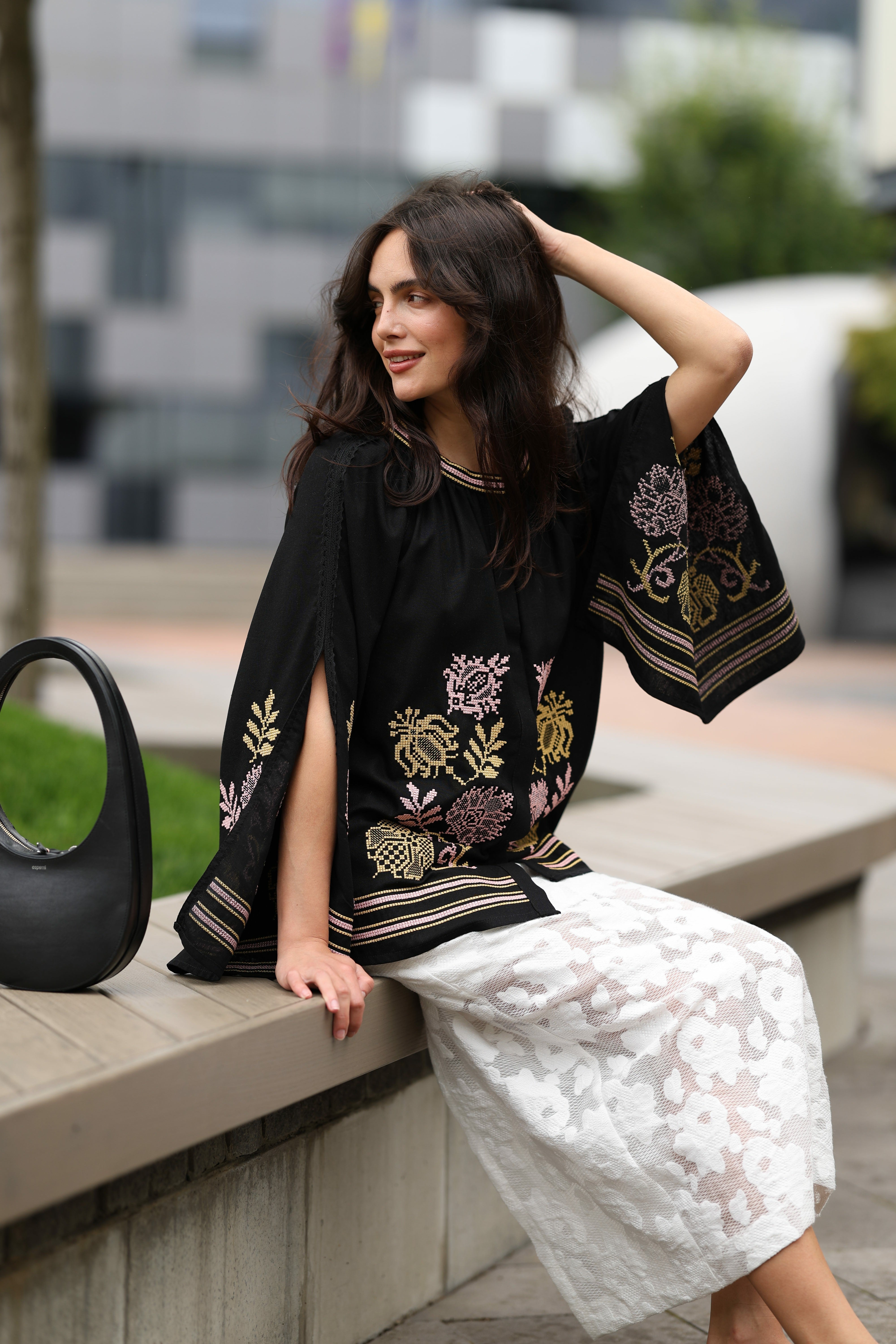 Woman sitting on a bench wearing a black embroidered top and white lace skirt.