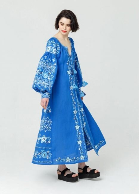 A woman modeling a long blue embroidered dress with floral patterns and long sleeves.