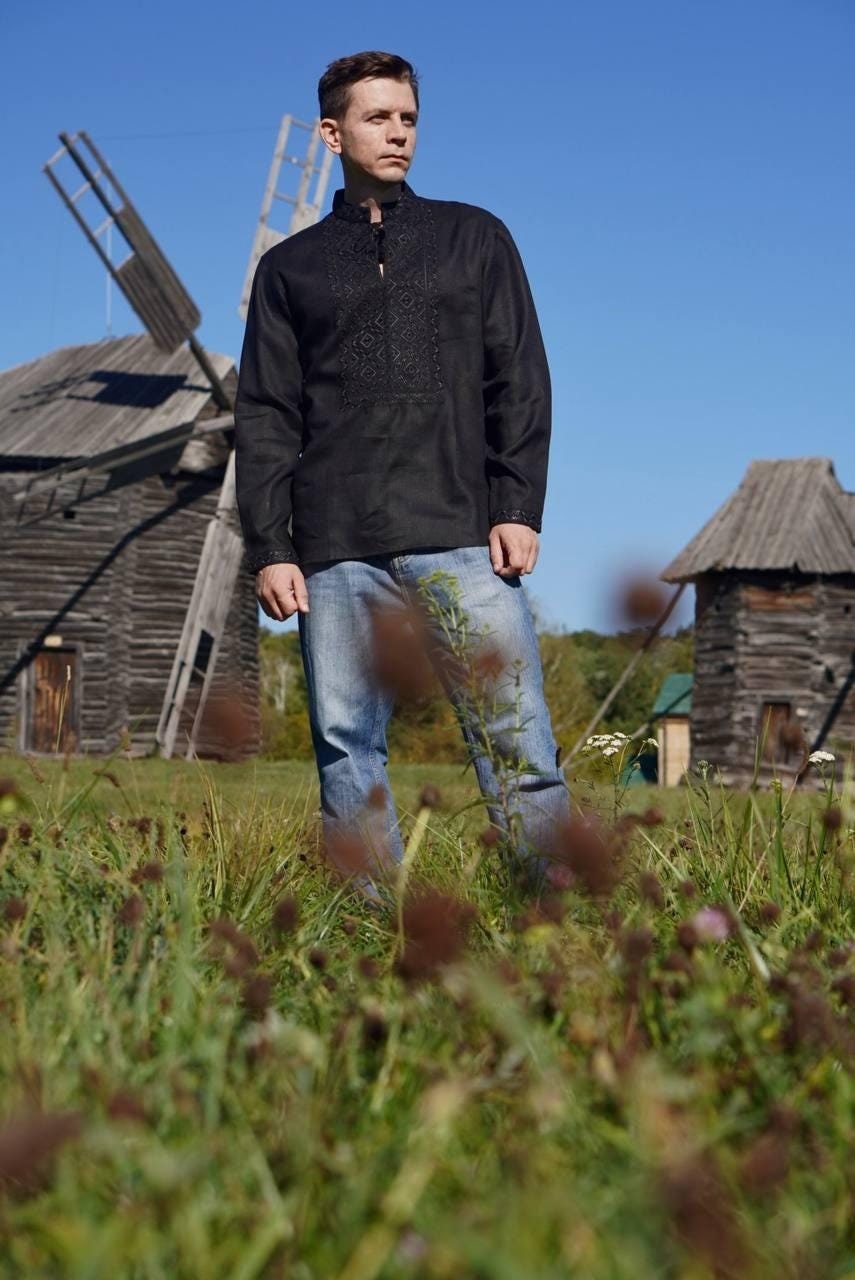 A man wearing a black embroidered Ukrainian shirt with white patterns, standing in front of traditional wooden Ukrainian houses.