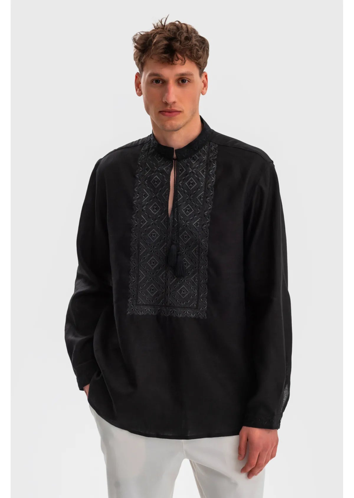 A man wearing a black long-sleeve kurta with a high collar and a dark embroidered geometric pattern on the chest, paired with white trousers, against a light gray background.