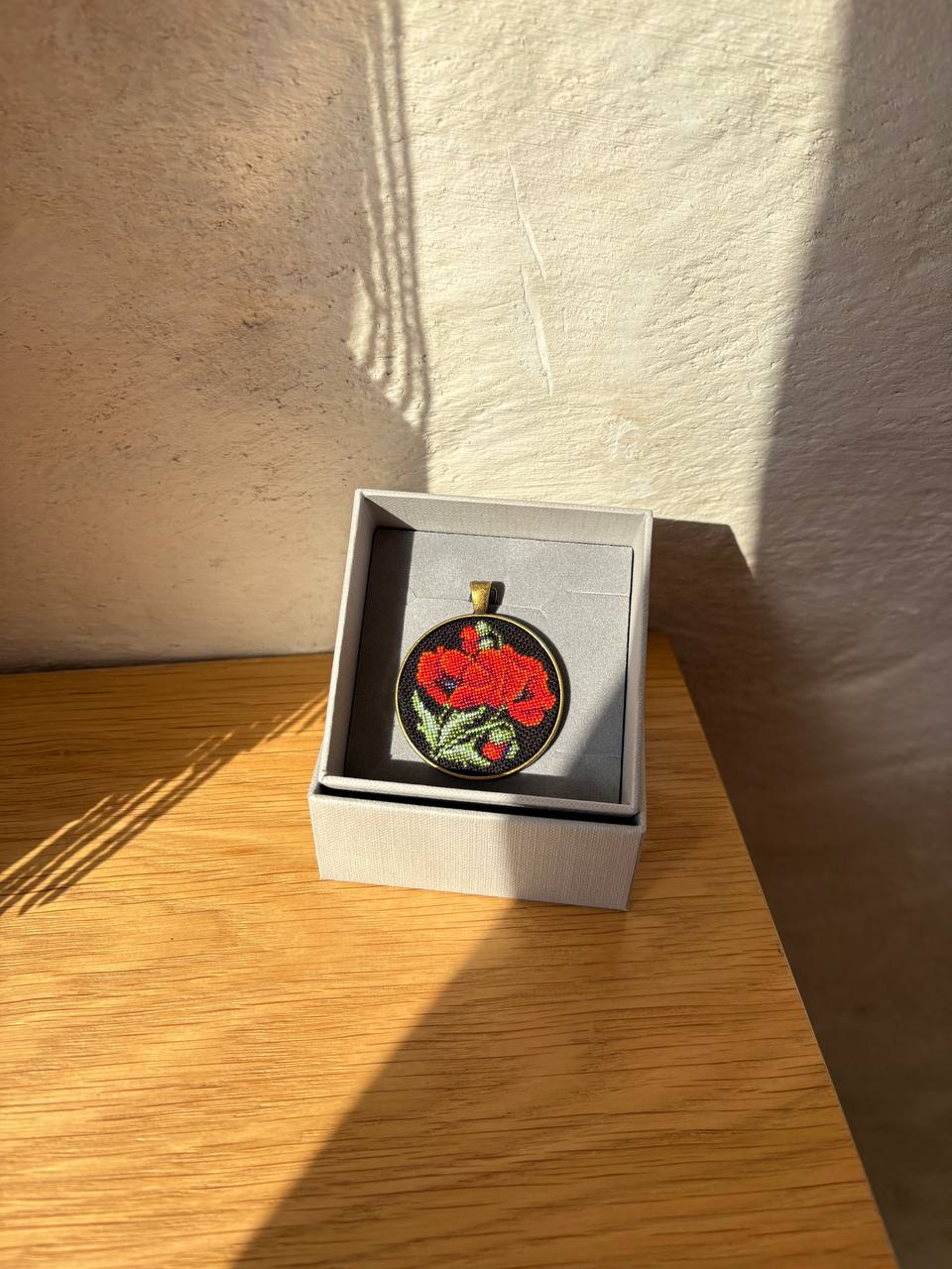 An oval pendant with a red floral design on a black background, displayed inside a light gray jewelry box on a wooden surface.